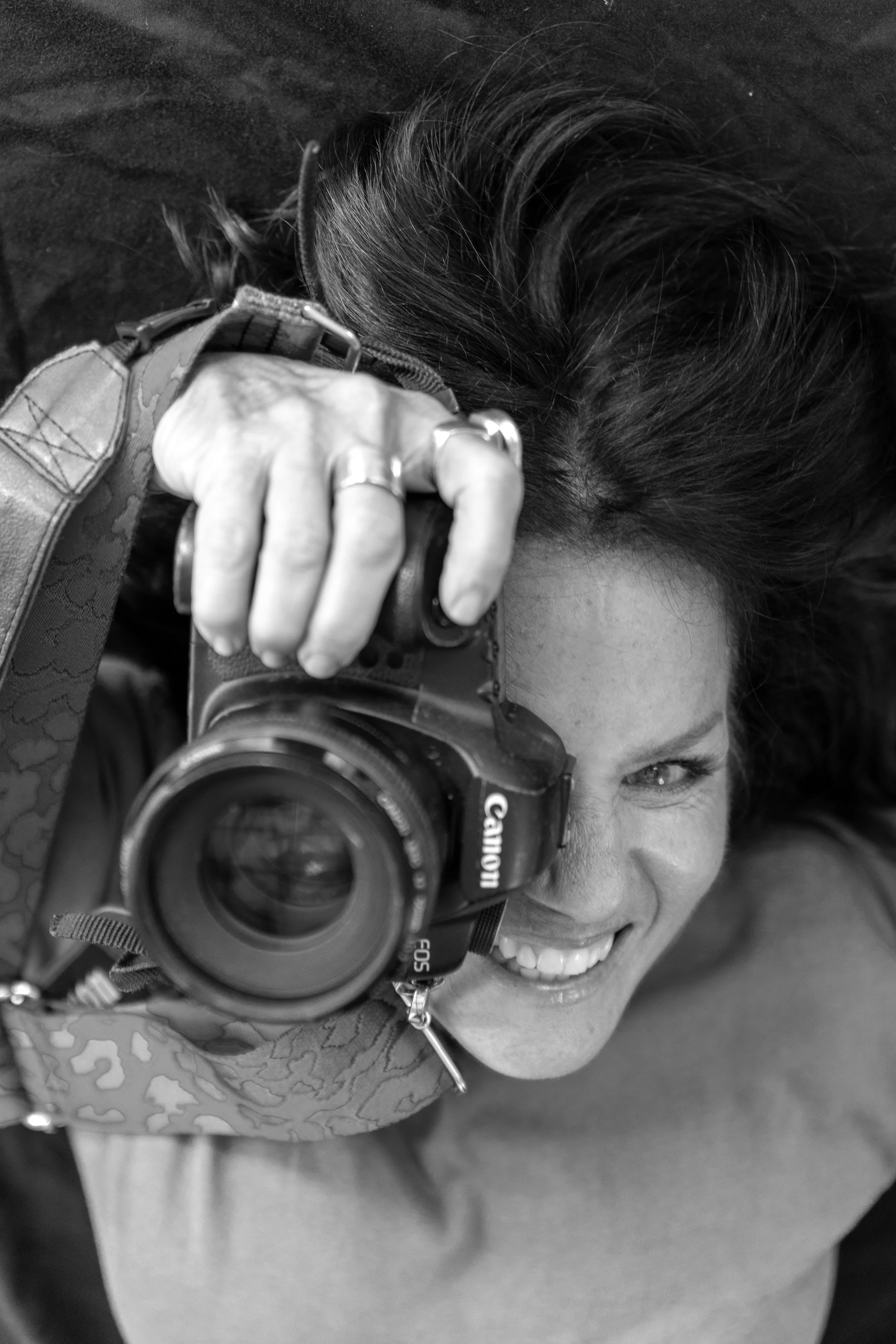 Black and white photo of a woman lying down, smiling, while holding a Canon DSLR camera up to her face, looking through the viewfinder.