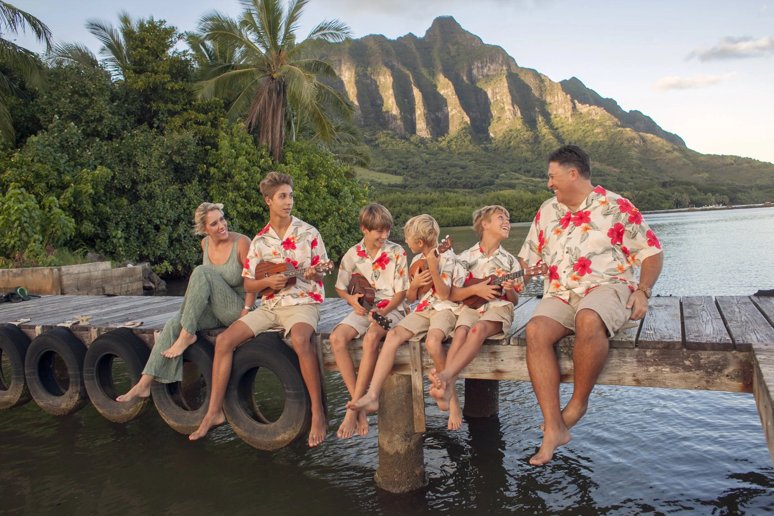 A family of seven sitting on a wooden dock by the water, playing ukuleles and smiling, with tropical trees and mountains in the background, all dressed in matching Hawaiian shirts.