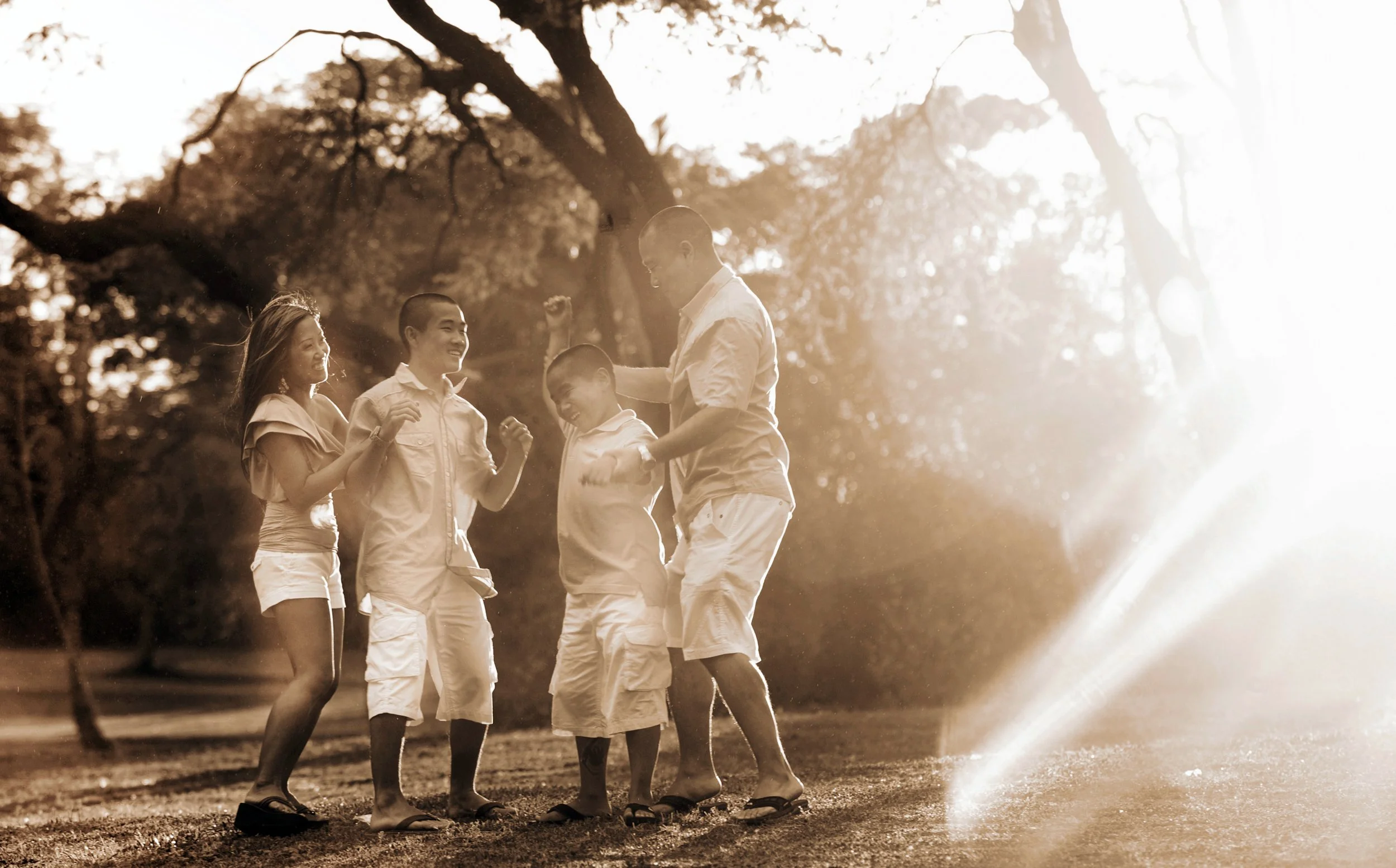 A family of five, including children and a parent, enjoying fun in a park during sunset with a water sprinkler, smiling and playing together.