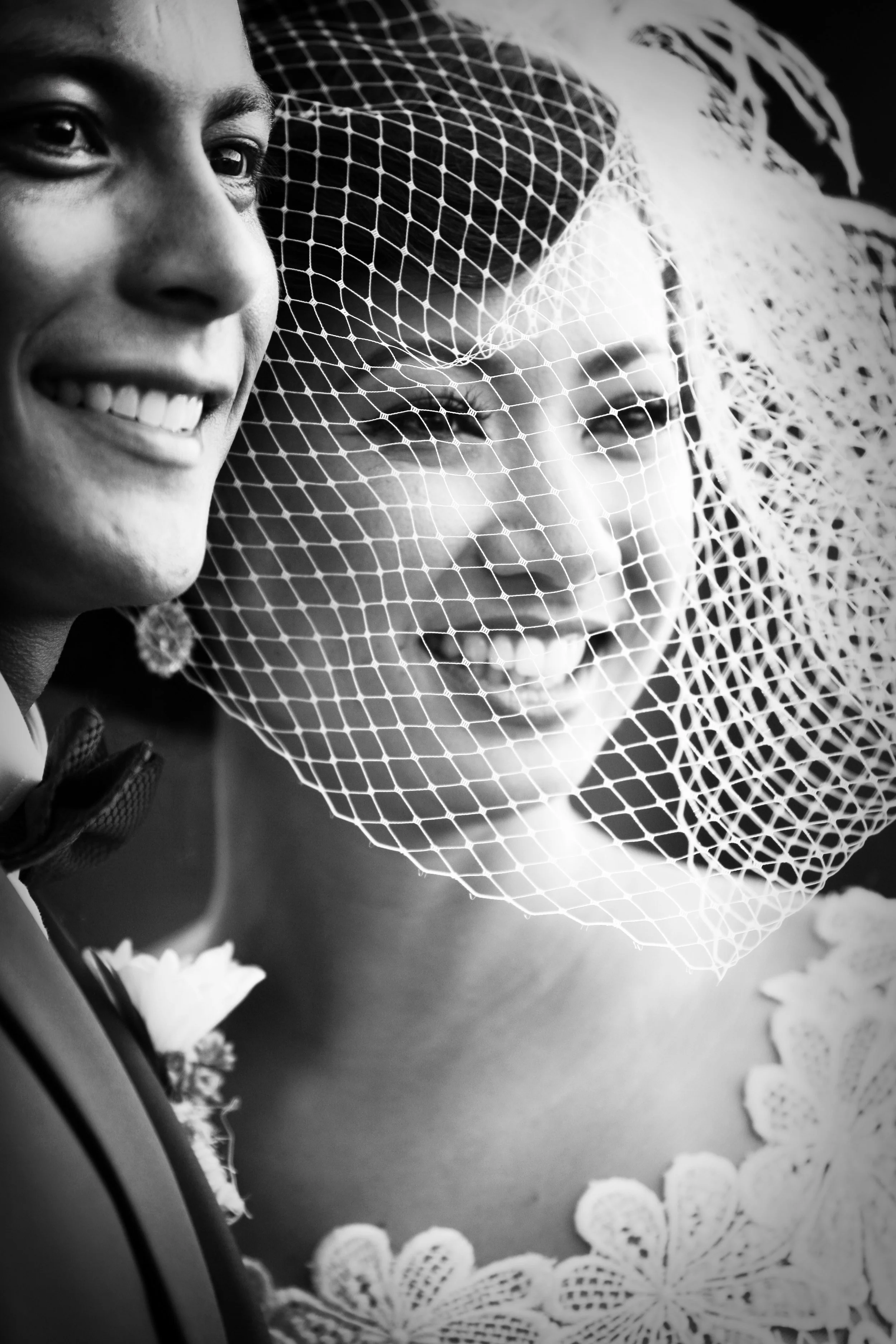 Black and white photo of a smiling man and a woman wearing a veil, lace dress, and floral accessory at a wedding.