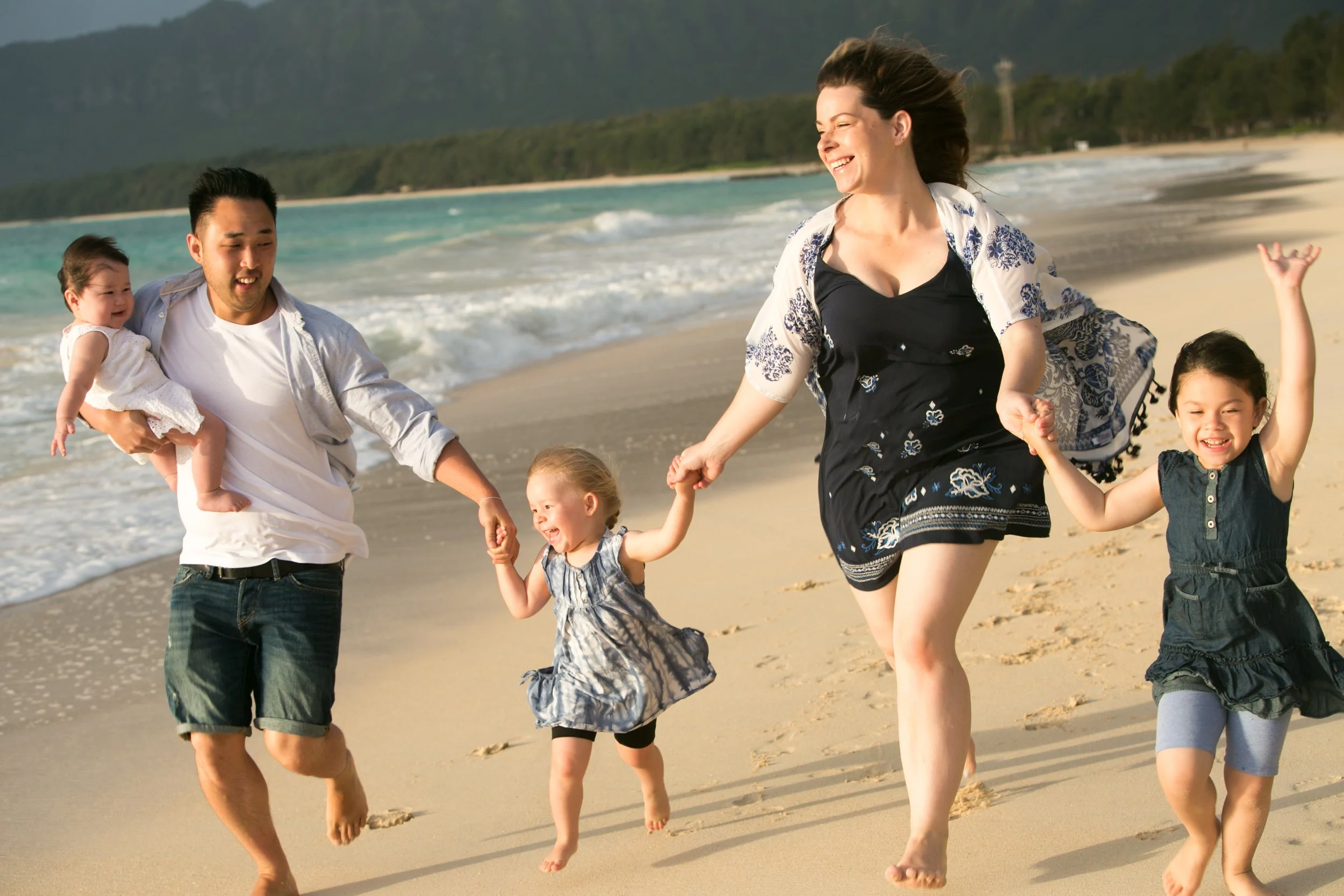 A family of five, including two adults and three children, holding hands and running along the beach near the ocean, with mountains and trees in the background.