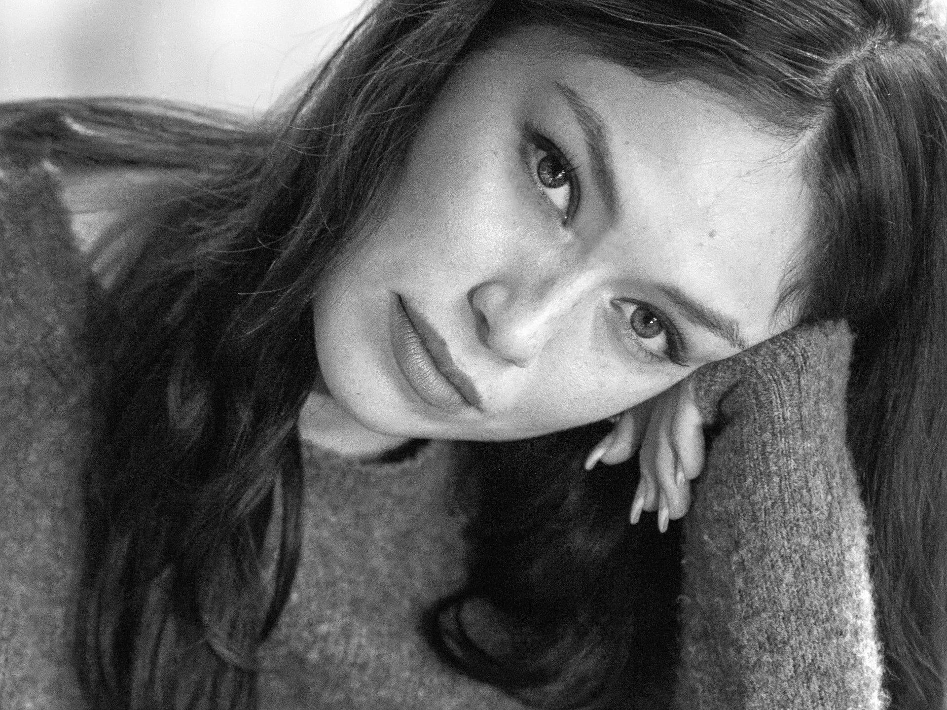 A black and white portrait of a woman with long dark hair, resting her head on her hand, looking into the camera.