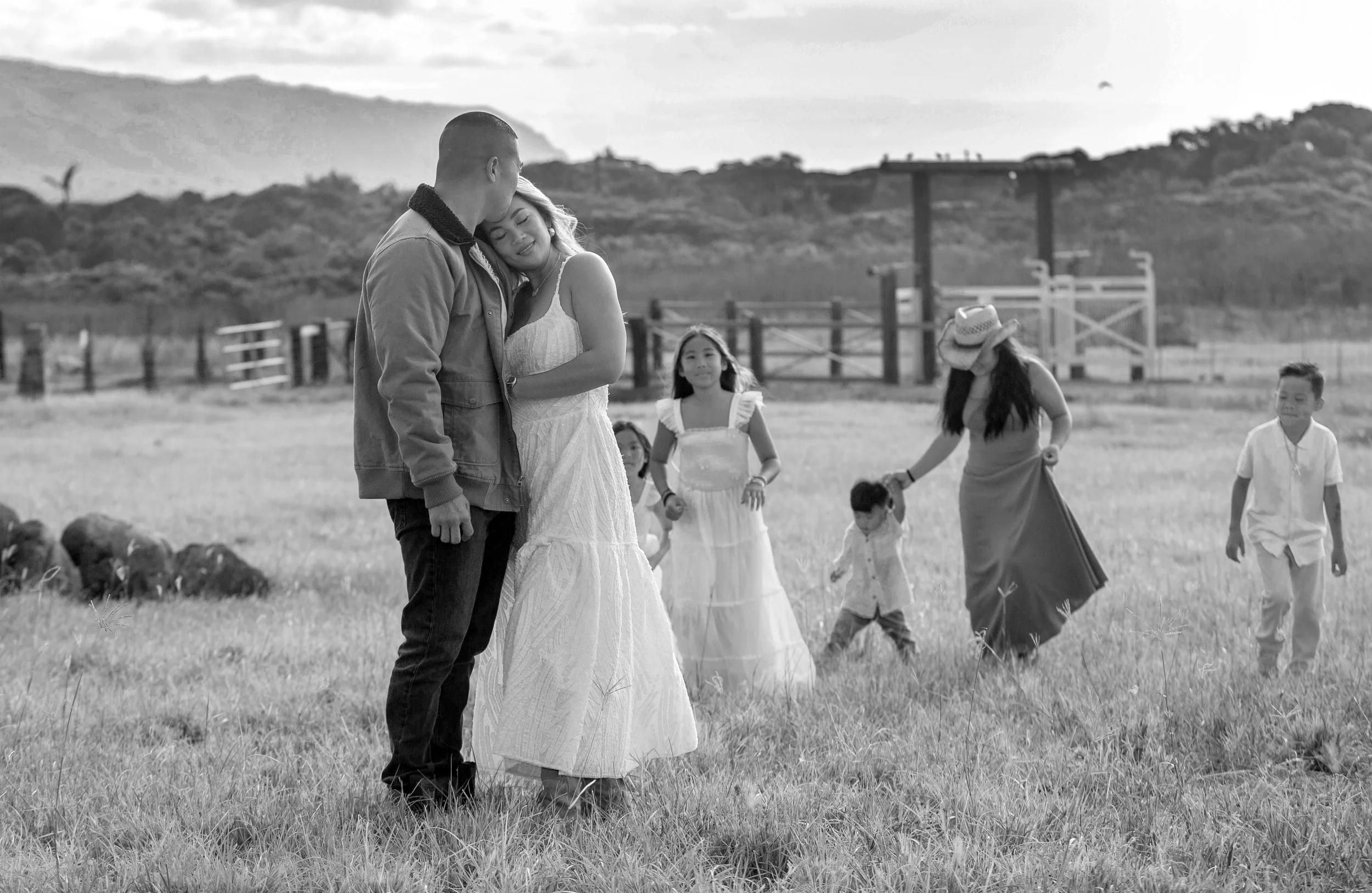 A couple embracing in a grassy field with children playing and a woman in a hat beside them, with a fence and hills in the background.
