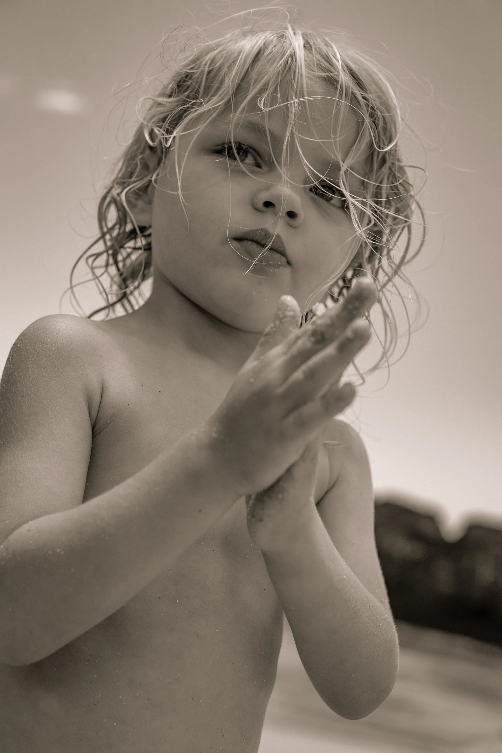 A young girl with wet, curly hair looks contemplative while holding her hands together in front of her chest. She is topless, with a blurred outdoor background.