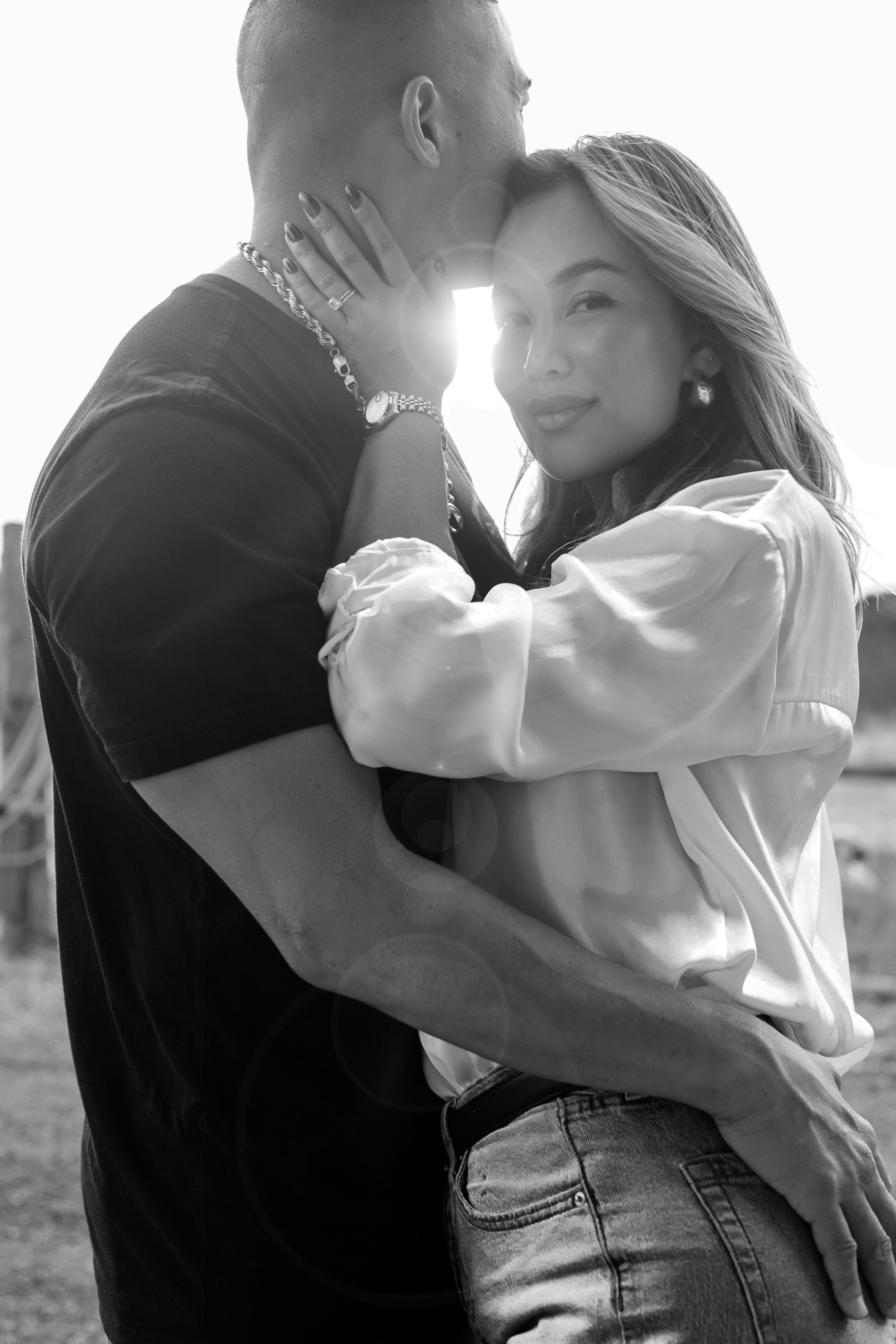 A black and white photo of a couple embracing outdoors, with the woman smiling directly at the camera and the man kissing her on the forehead. The woman has long hair, earrings, and a satin shirt, while the man has short hair, a black t-shirt, and is