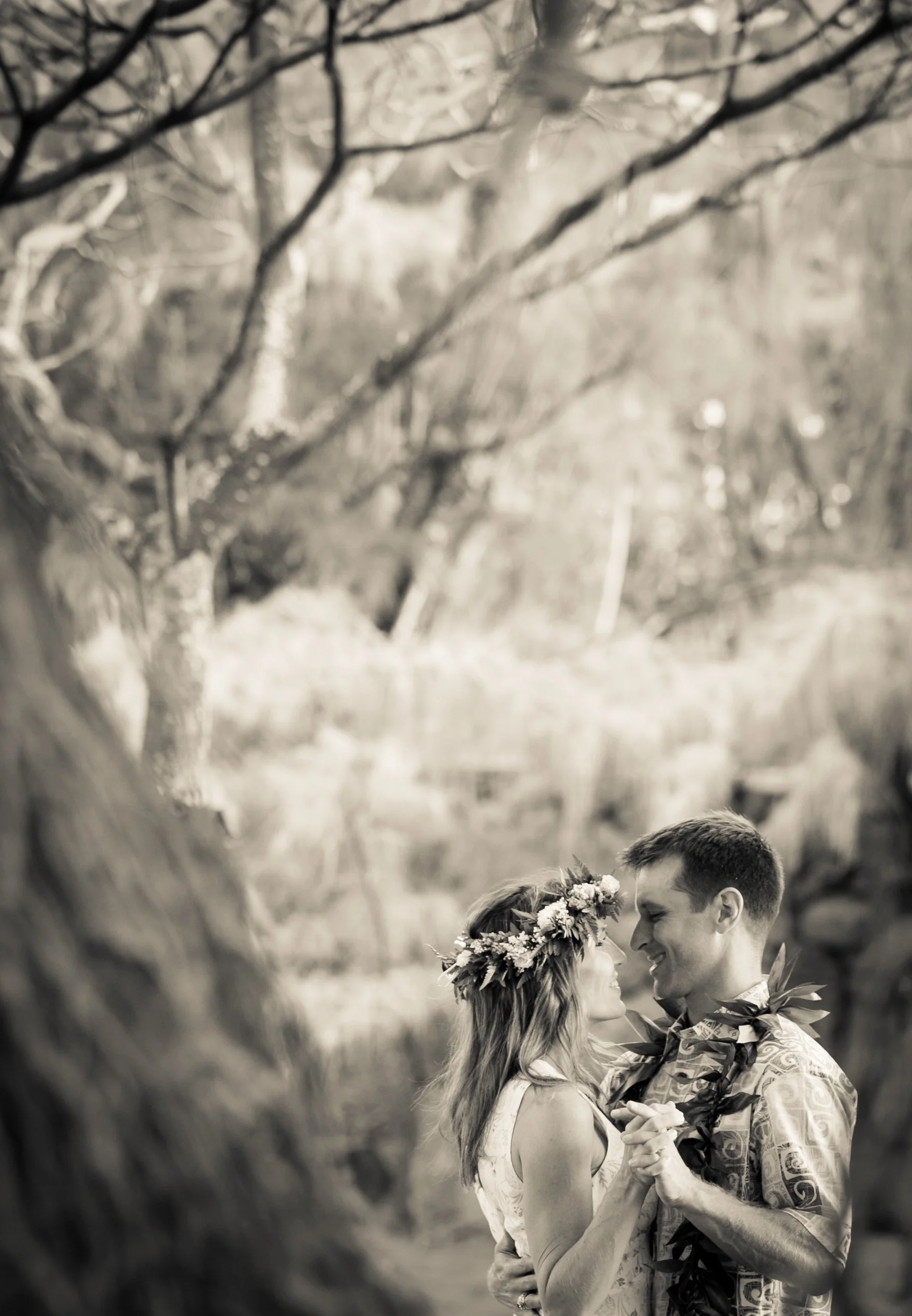 A couple dancing outdoors, decorated with leis and a floral crown, surrounded by trees.