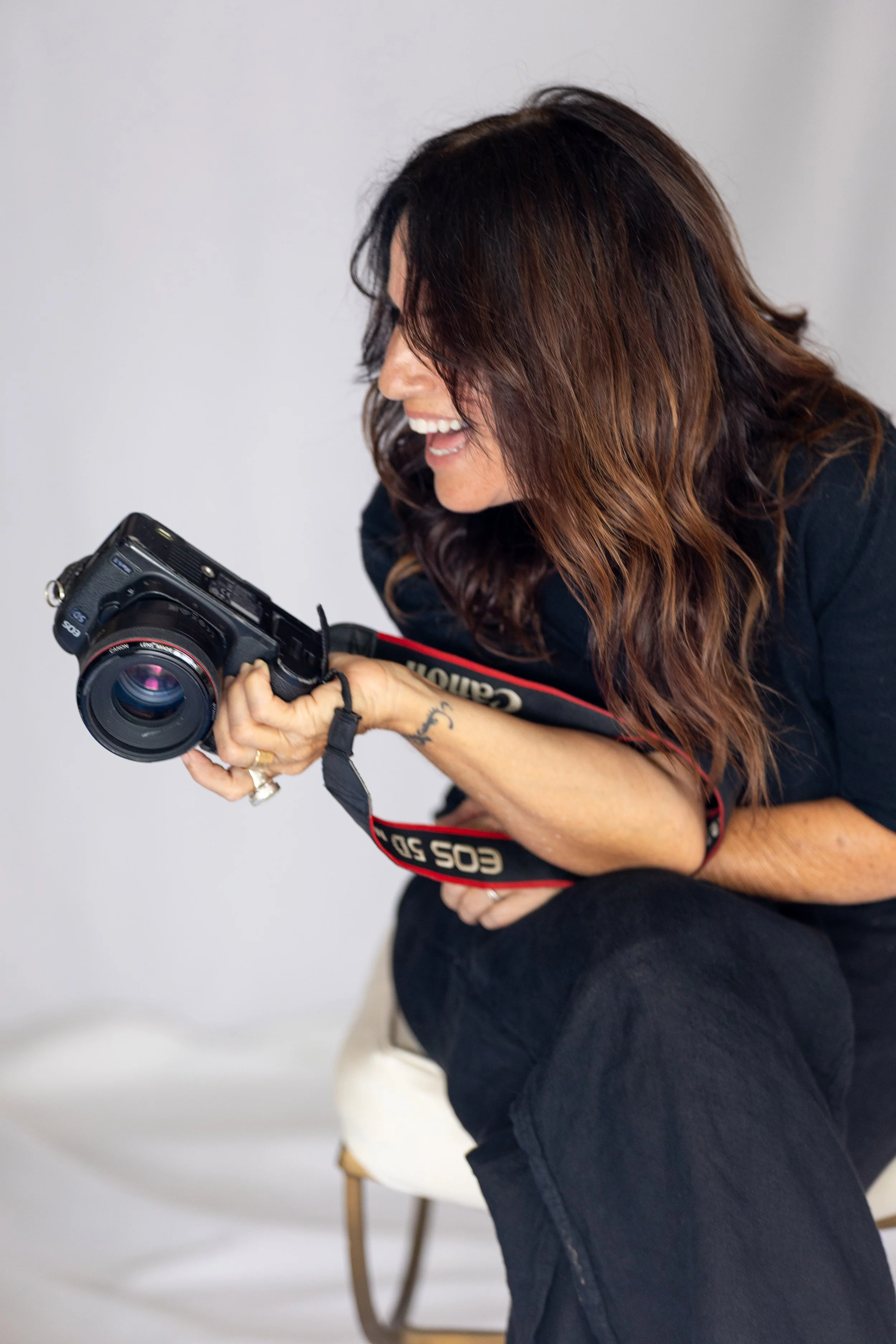 A woman with brown hair holding a Canon camera, sitting on a white chair, smiling and looking down at her camera.