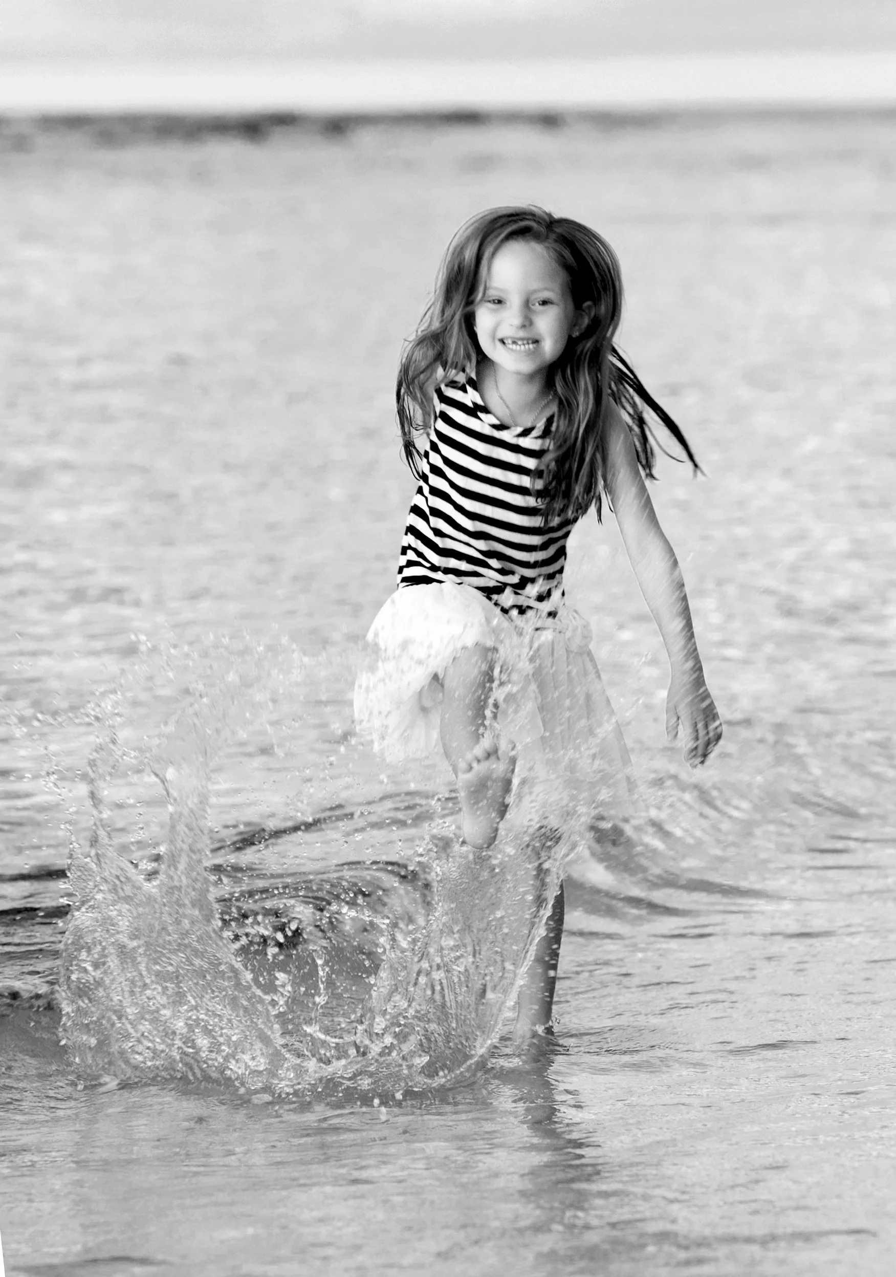 A young girl playing and running through the shallow water at the beach, smiling and splashing.