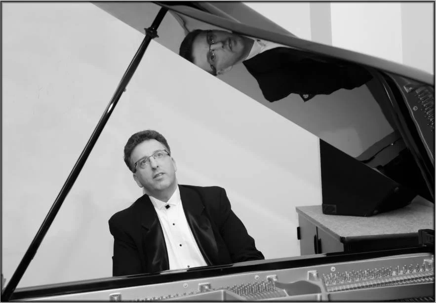 A black and white photo of two men, one sitting at a grand piano and the other looking over the edge of the piano, with their reflections visible on the piano's lid.