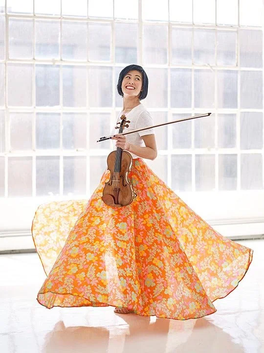 A woman standing in front of a large window, holding a violin and bow, wearing a white top and a long, flowing orange and yellow floral skirt.