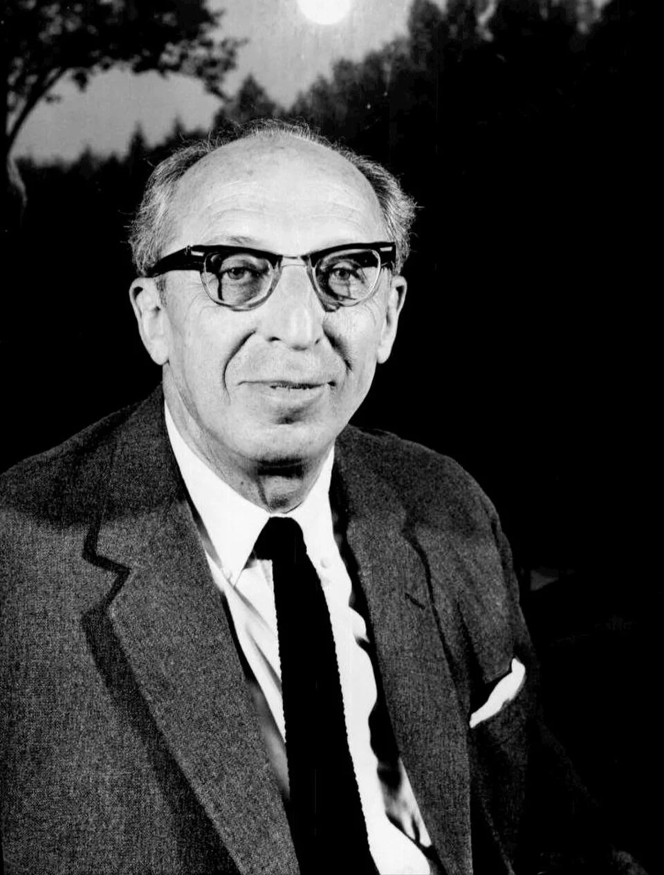 Black and white photo of an older man wearing glasses, a suit, and a tie, outdoors with trees and a bright sky in the background.