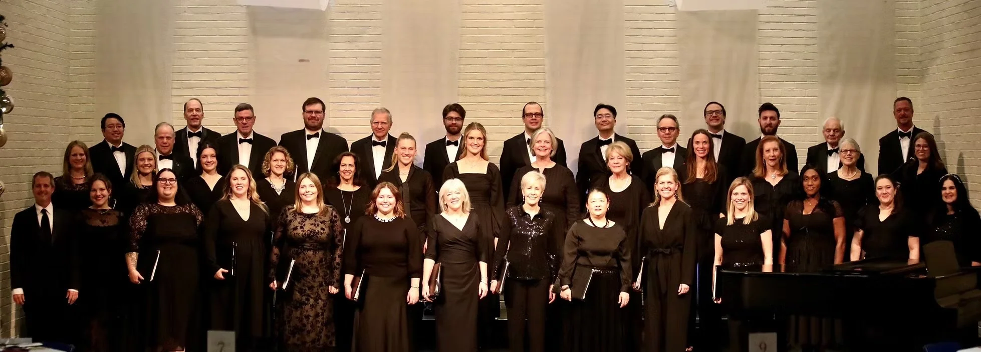 Group photo of men and women in formal attire, standing on risers in front of a brick wall, for a choir or musical performance.