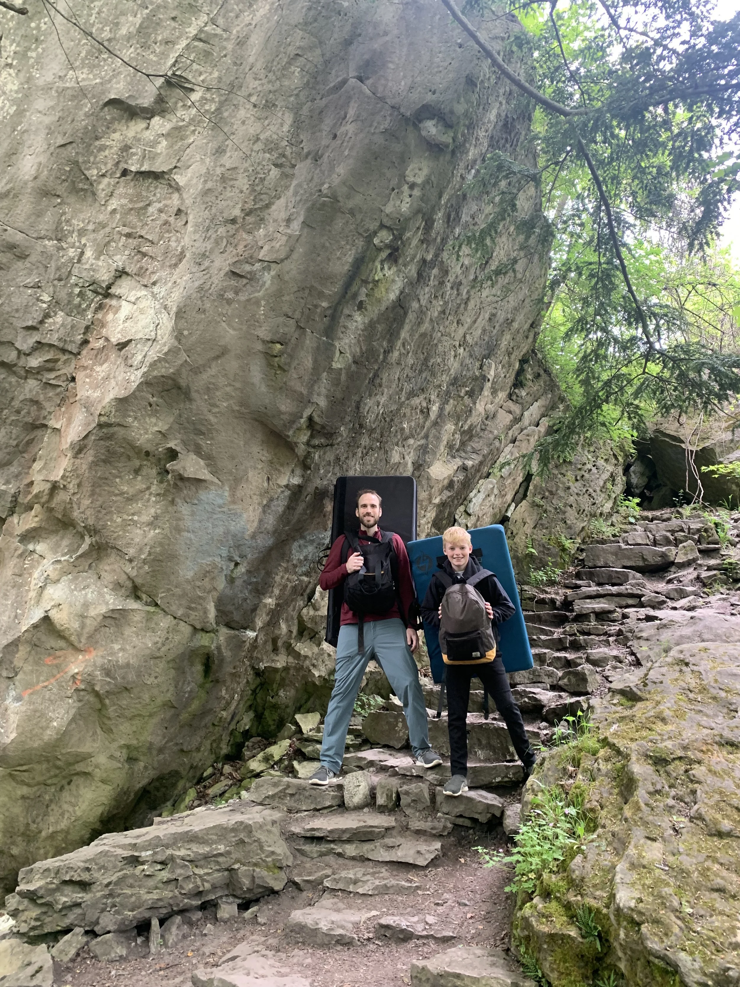 Two men with climbing gear and backpacks standing on a rocky trail in a forest near large rock formations and stone steps.