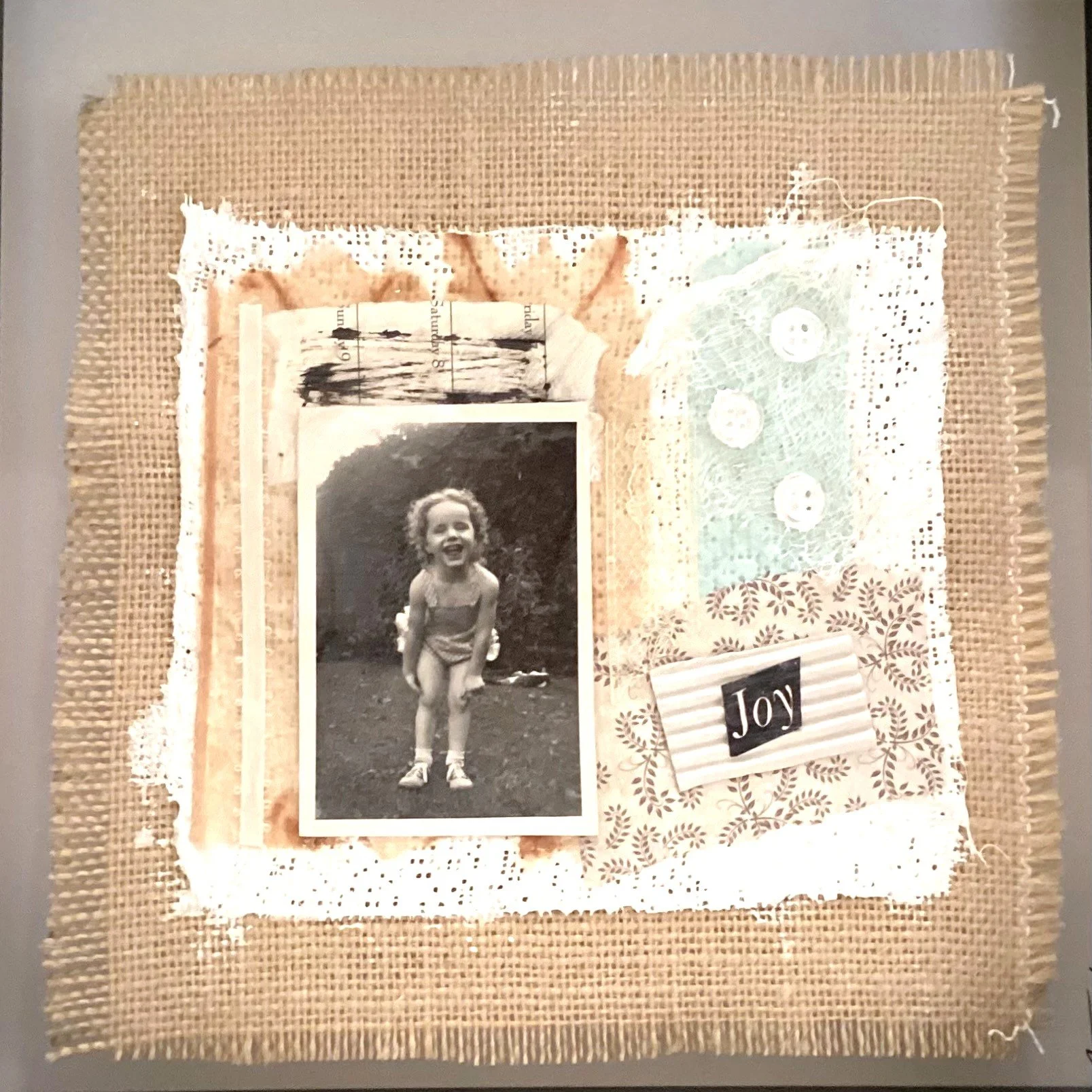 A framed black and white photo of a smiling young girl, standing outdoors on grass, wearing a sleeveless top and shorts, surrounded by fabric, lace, and decorative paper with a 'Joy' label.
