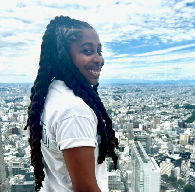 Smiling woman with long dreadlocks on a rooftop, overlooking a city skyline and cloudy sky.