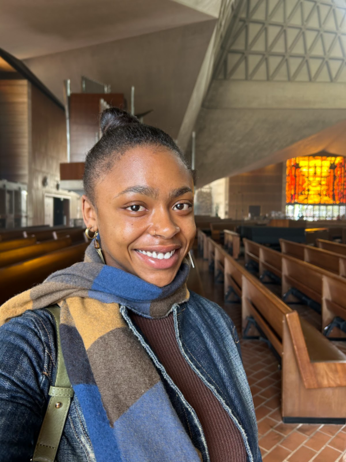A young woman takes a selfie inside a church, smiling, with wooden pews and colorful stained glass window in the background.
