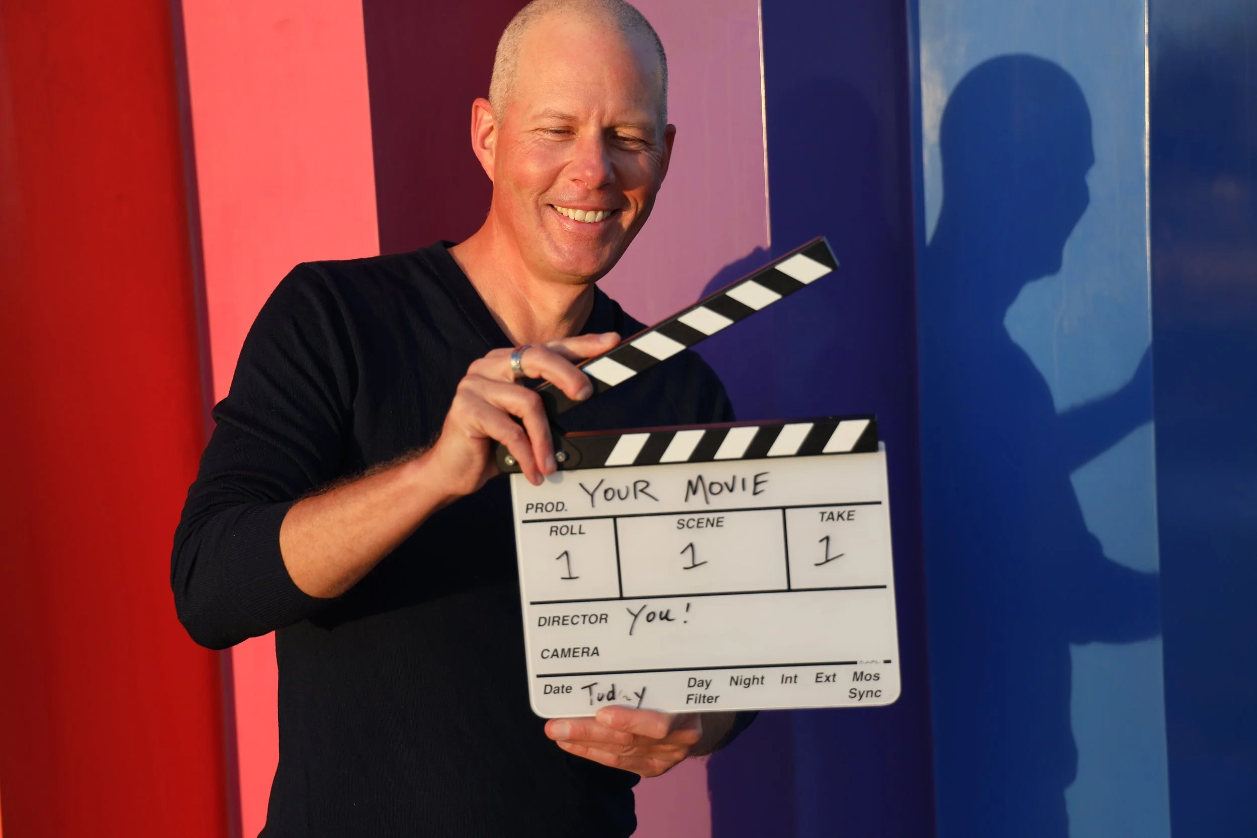 A smiling man holding a film slate that reads 'YOUR MOVIE' with the scene number 1, take 1, director 'You!', and the date 'Tuesday'. He is standing against a colorful background with vertical sections of red, purple, and blue.