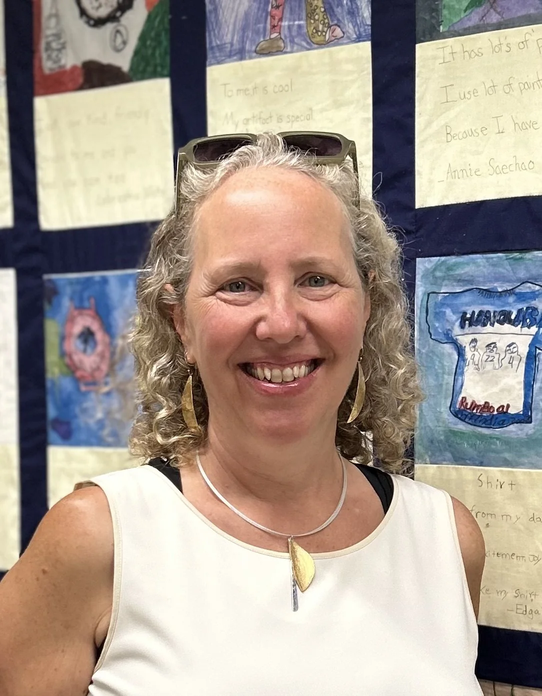 A woman with curly blonde hair wearing a white sleeveless top, earrings, and a necklace, smiling in front of a display of children's artwork.