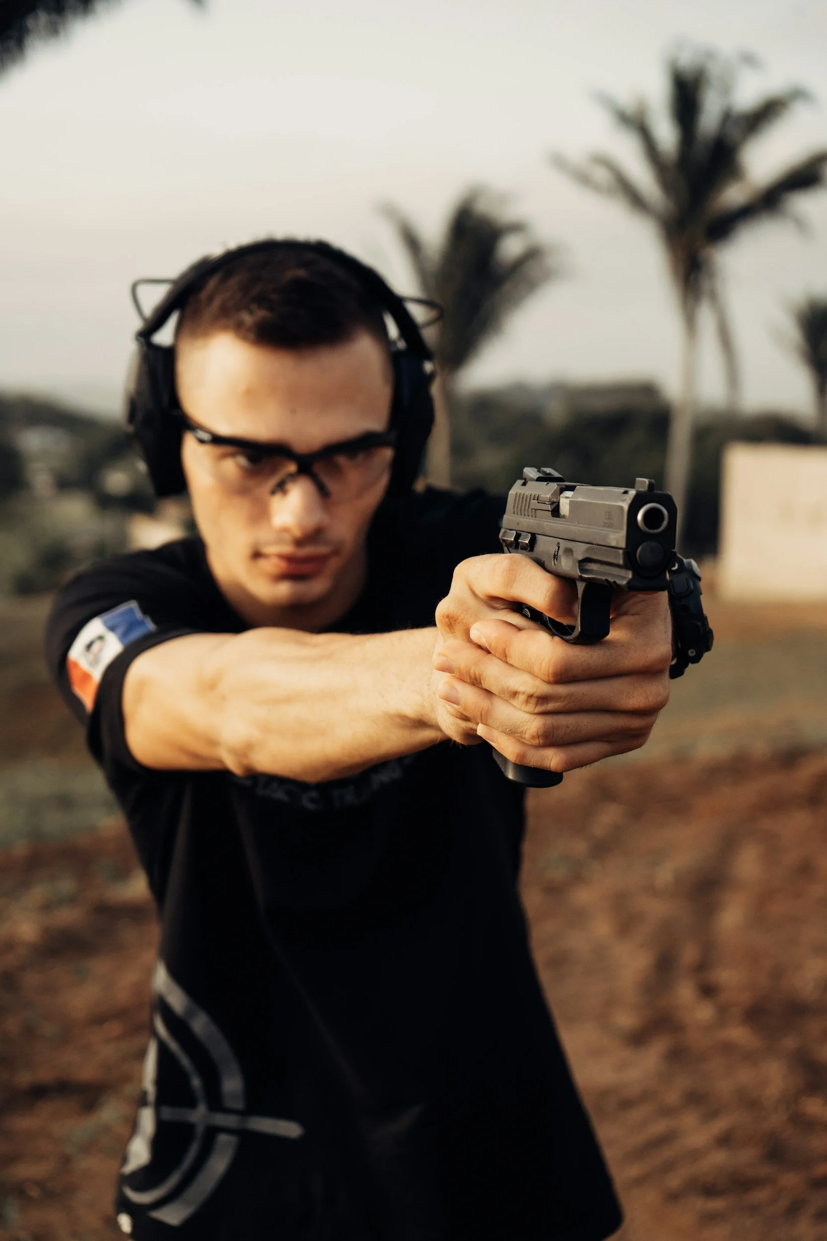 A man aiming a handgun outdoors, wearing protective glasses and ear protection, with palm trees and a cloudy sky in the background.