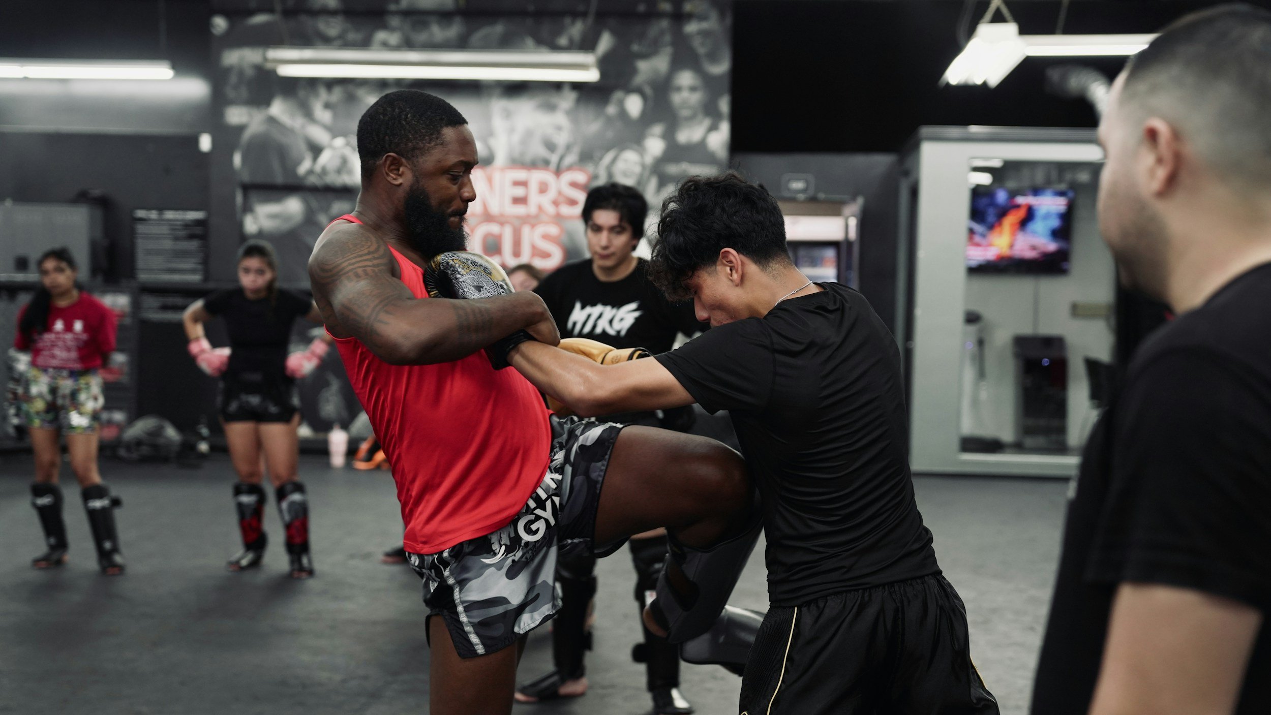 A mixed martial arts fighter in red shorts and a black t-shirt practicing knee strikes with a trainer in a gym. Other fighters and trainers observe in the background.