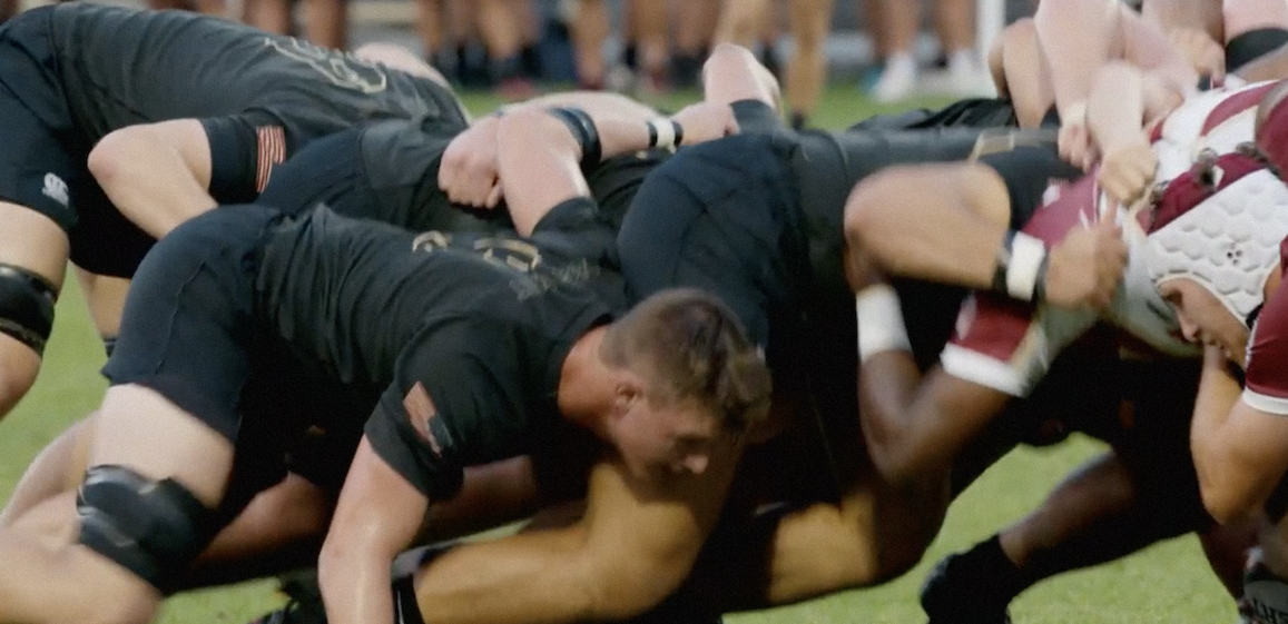A still from 'Brothers On Three'. A group of rugby players in black uniforms huddled together during a game.