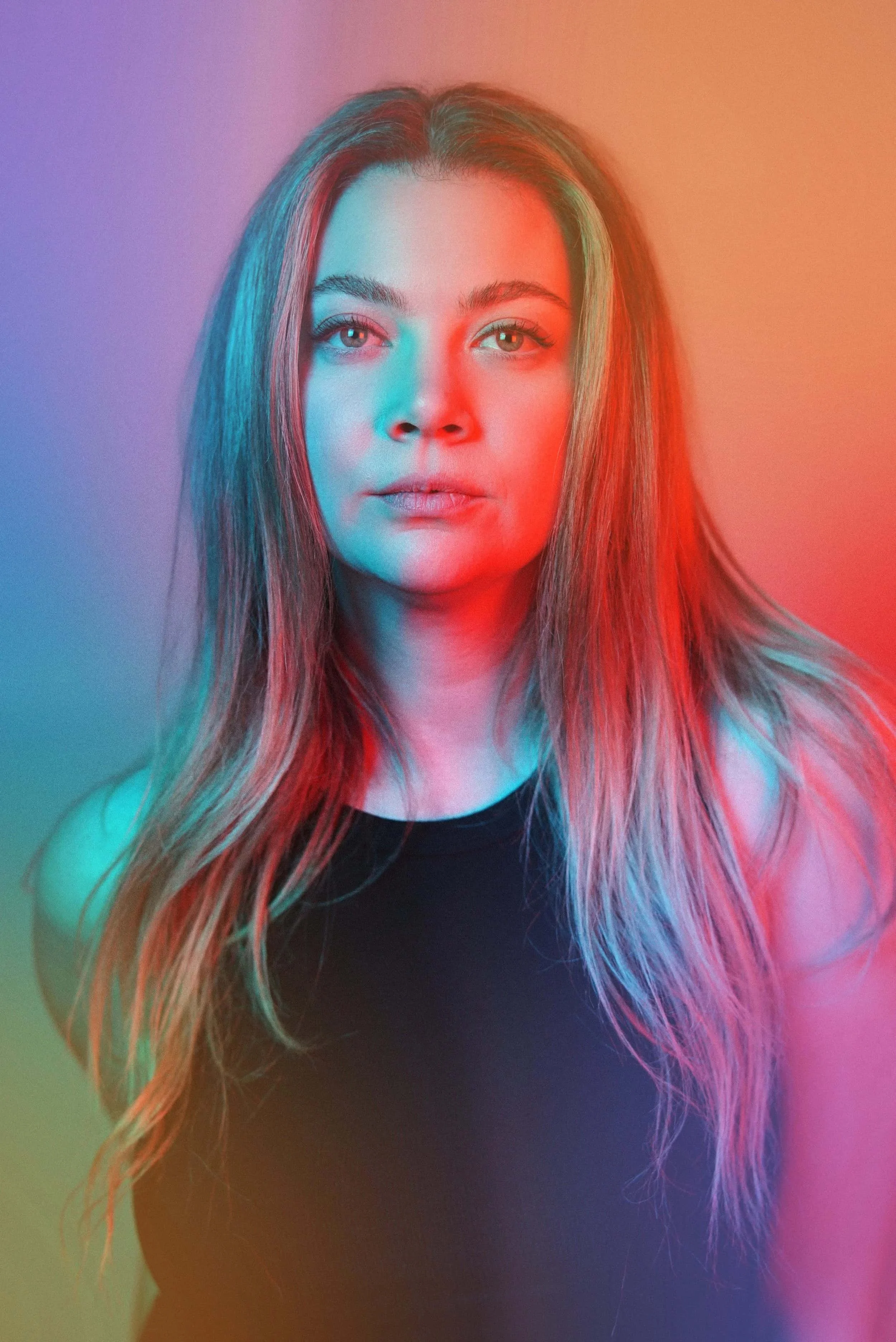 Portrait of a Rachel A Weise with long, wavy hair, wearing a black top, illuminated by colorful lighting in blue, red, yellow, and purple hues.