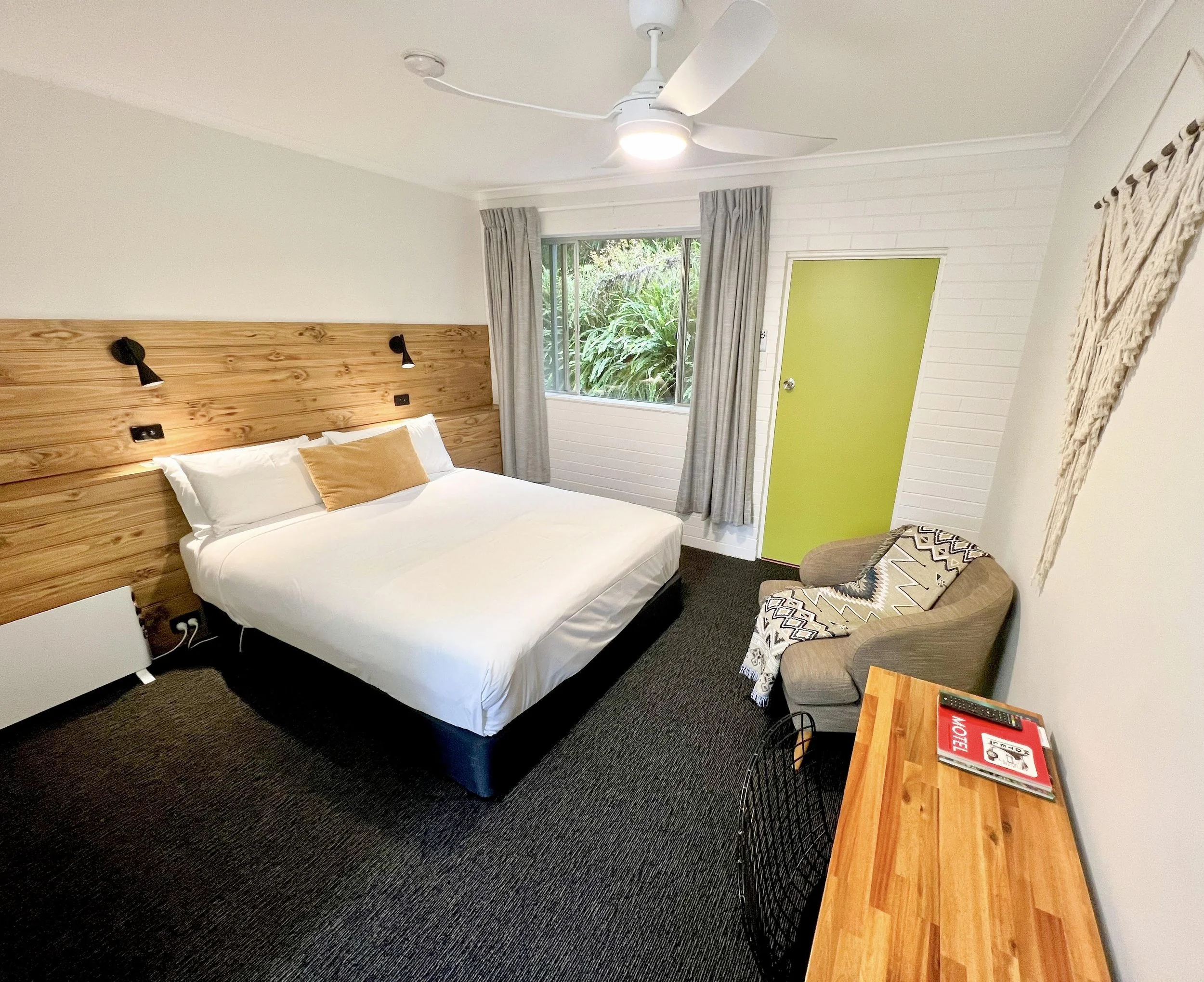 A bedroom with a bed, wooden headboard, and two small black reading lamps. There is a window with beige curtains, a bright green door, a patterned chair, and a wooden side table with magazines and a TV remote.