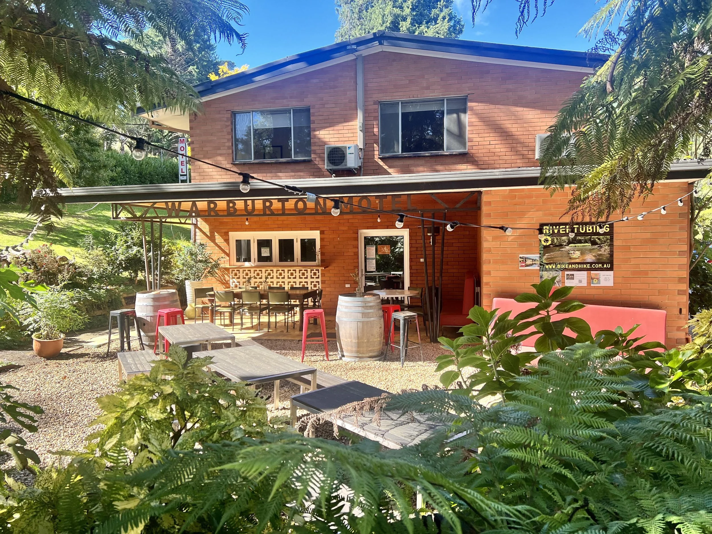 A two-story brick motel with a sign reading 'WARBURTON MOTEL', outdoor seating area with tables and chairs, surrounded by lush greenery and trees, with string lights hanging overhead.