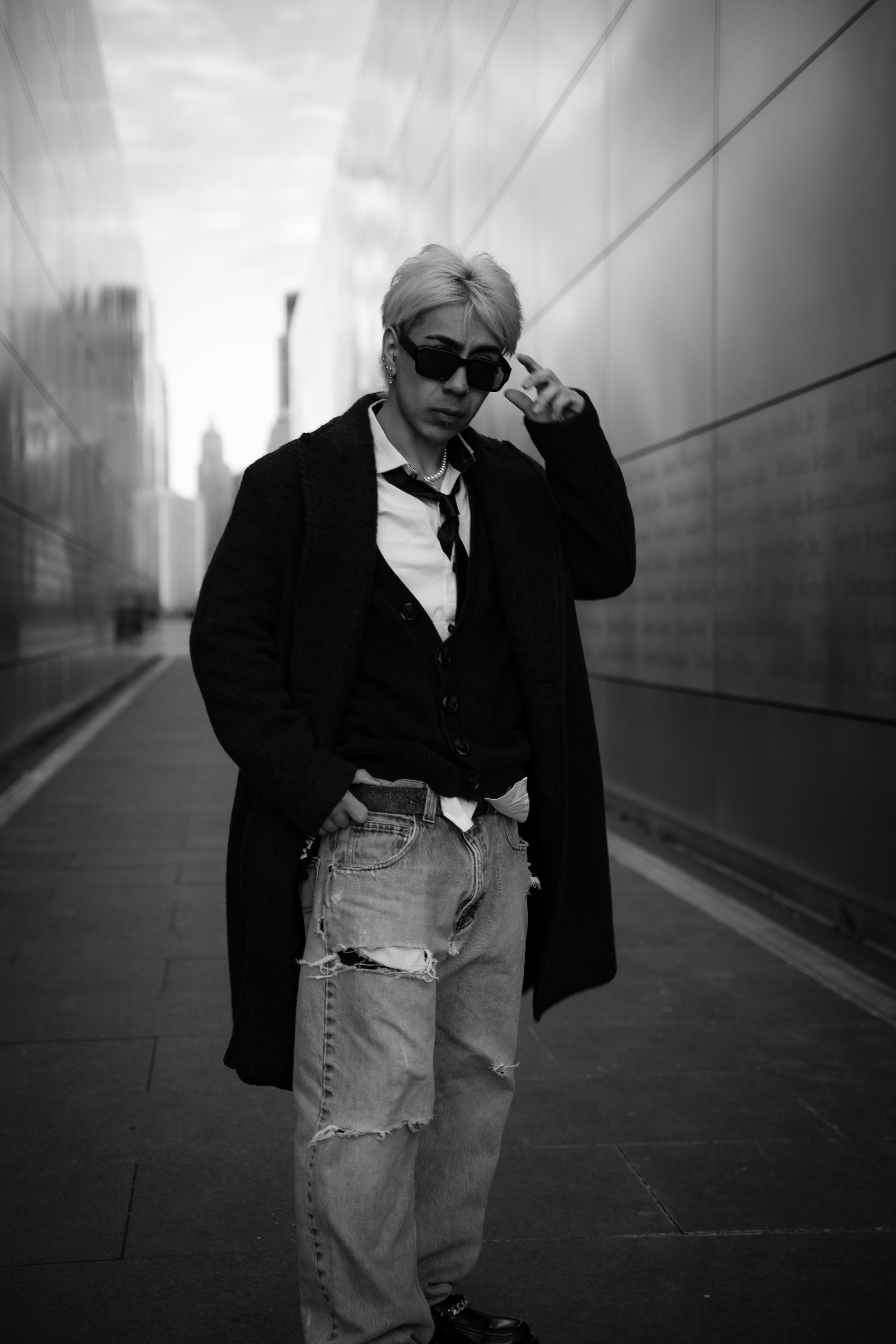 A woman with short blond hair wearing black sunglasses, a white shirt, a black blazer, a tie, a long dark coat, ripped jeans, and boots, standing on an urban sidewalk with modern buildings in the background.