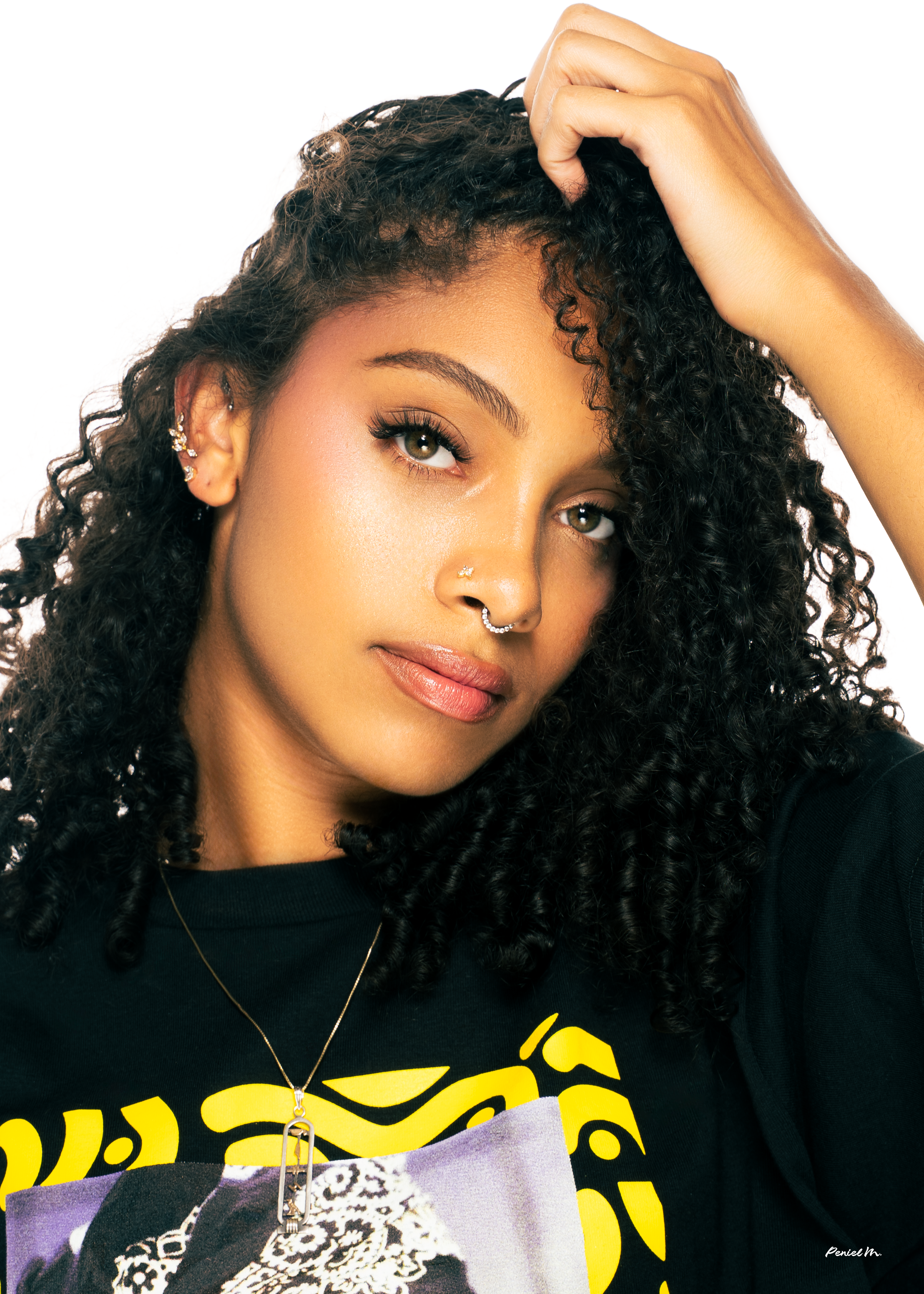 A woman with curly dark hair, multiple earrings, a septum nose ring, and a small nose stud, looking at the camera with a confident expression, wearing a black t-shirt with yellow text and a graphic, against a white background.