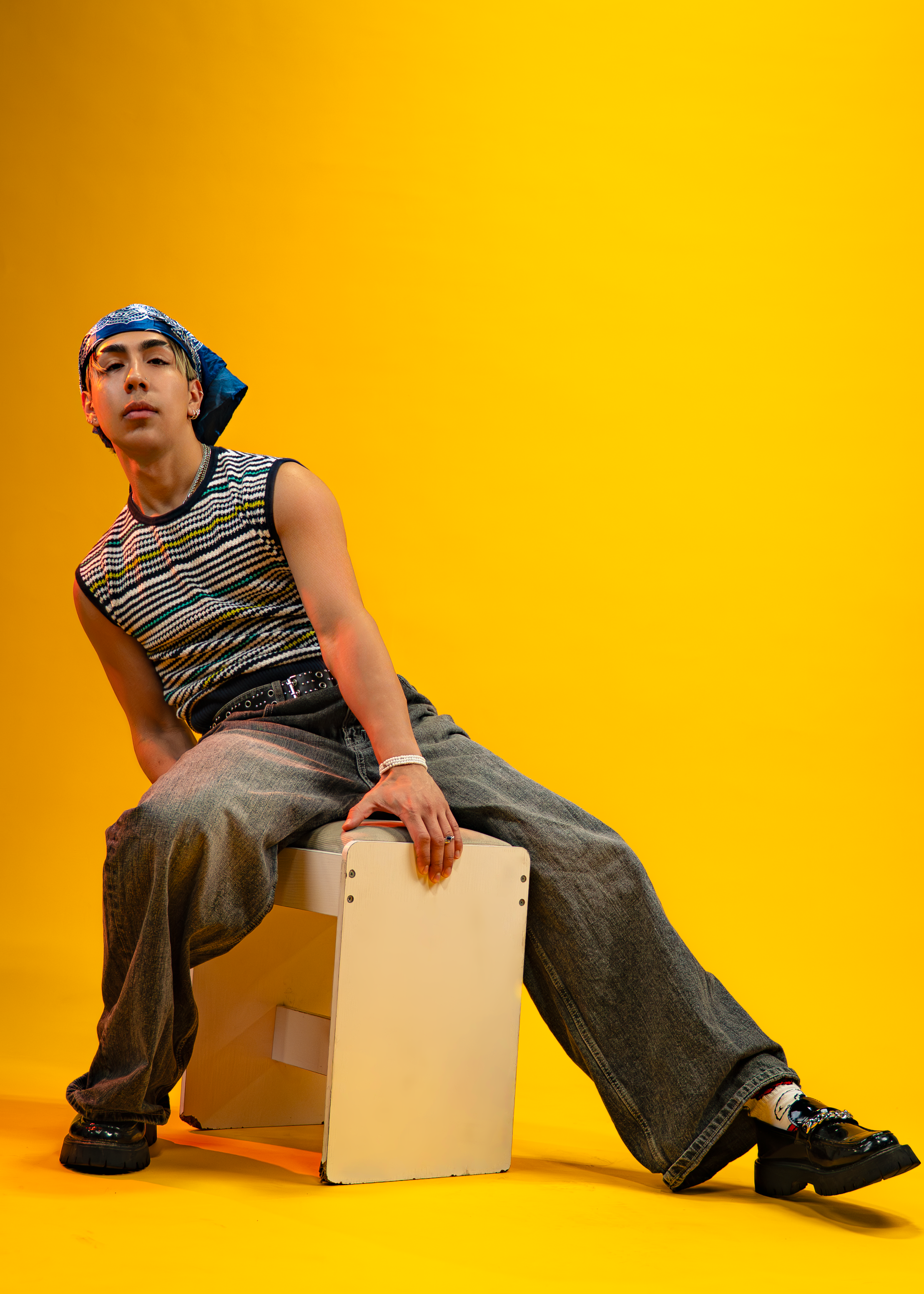 A person sitting on a white box against a yellow background, wearing a striped sleeveless top, baggy gray pants, black shoes with white socks, and a blue bandana on their head.