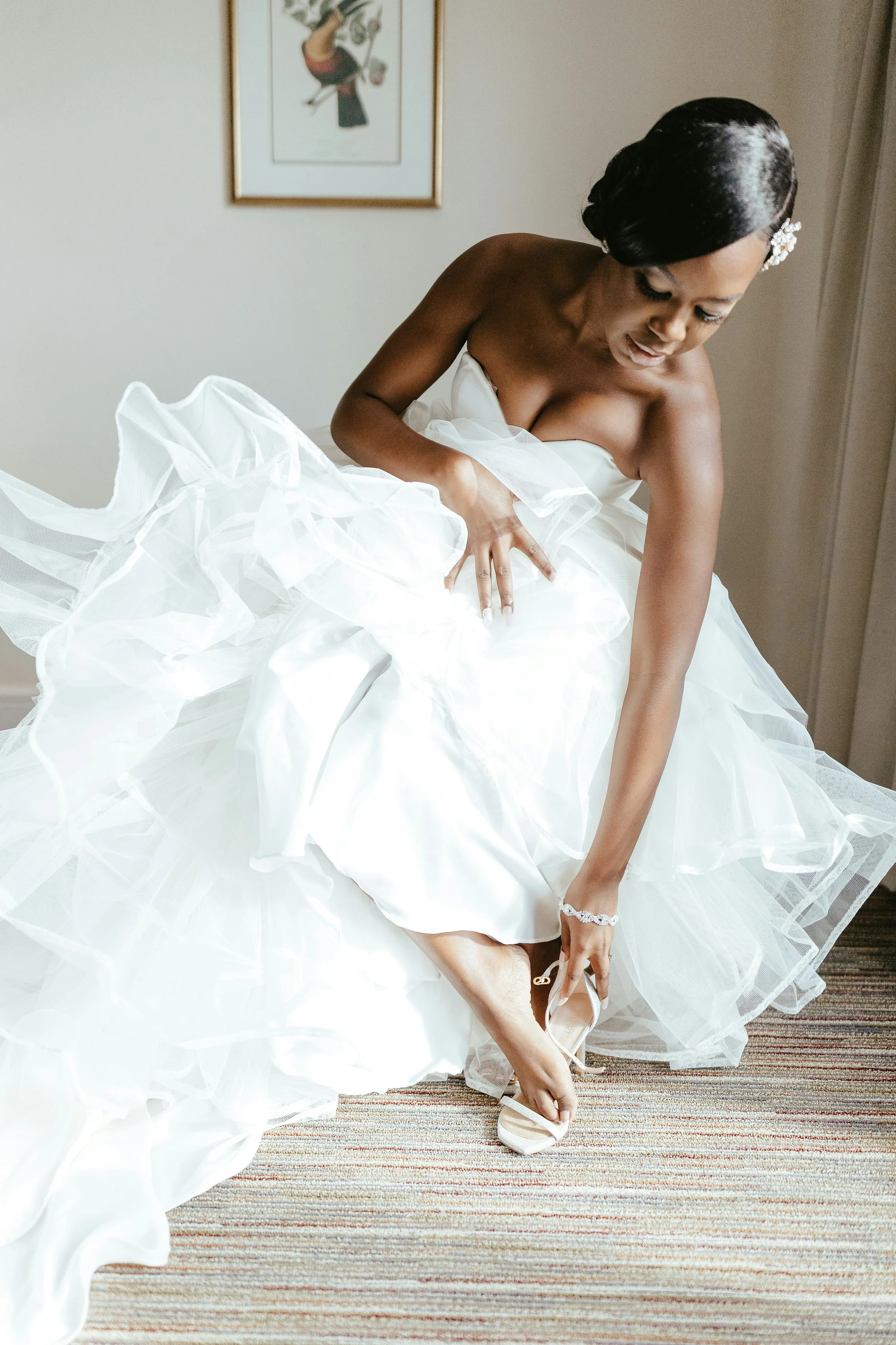 A bride in a white wedding dress is sitting on the floor, putting on her wedding shoes, in a softly lit room with a framed picture on the wall.