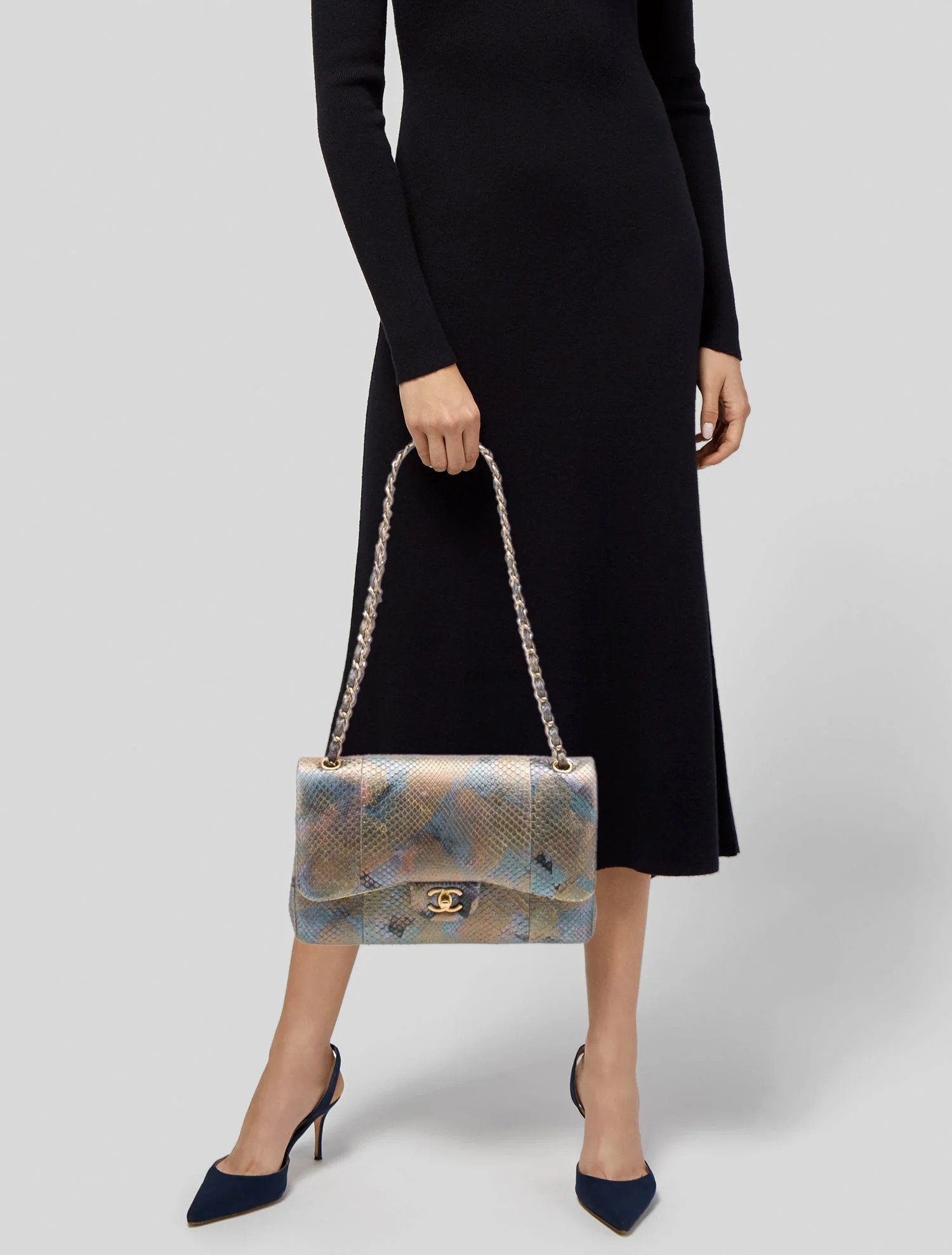 A woman wearing a black dress at mid-length, holding a colorful snakeskin-patterned handbag with a gold chain strap and logo, standing with one leg slightly in front of the other and wearing navy high heels.