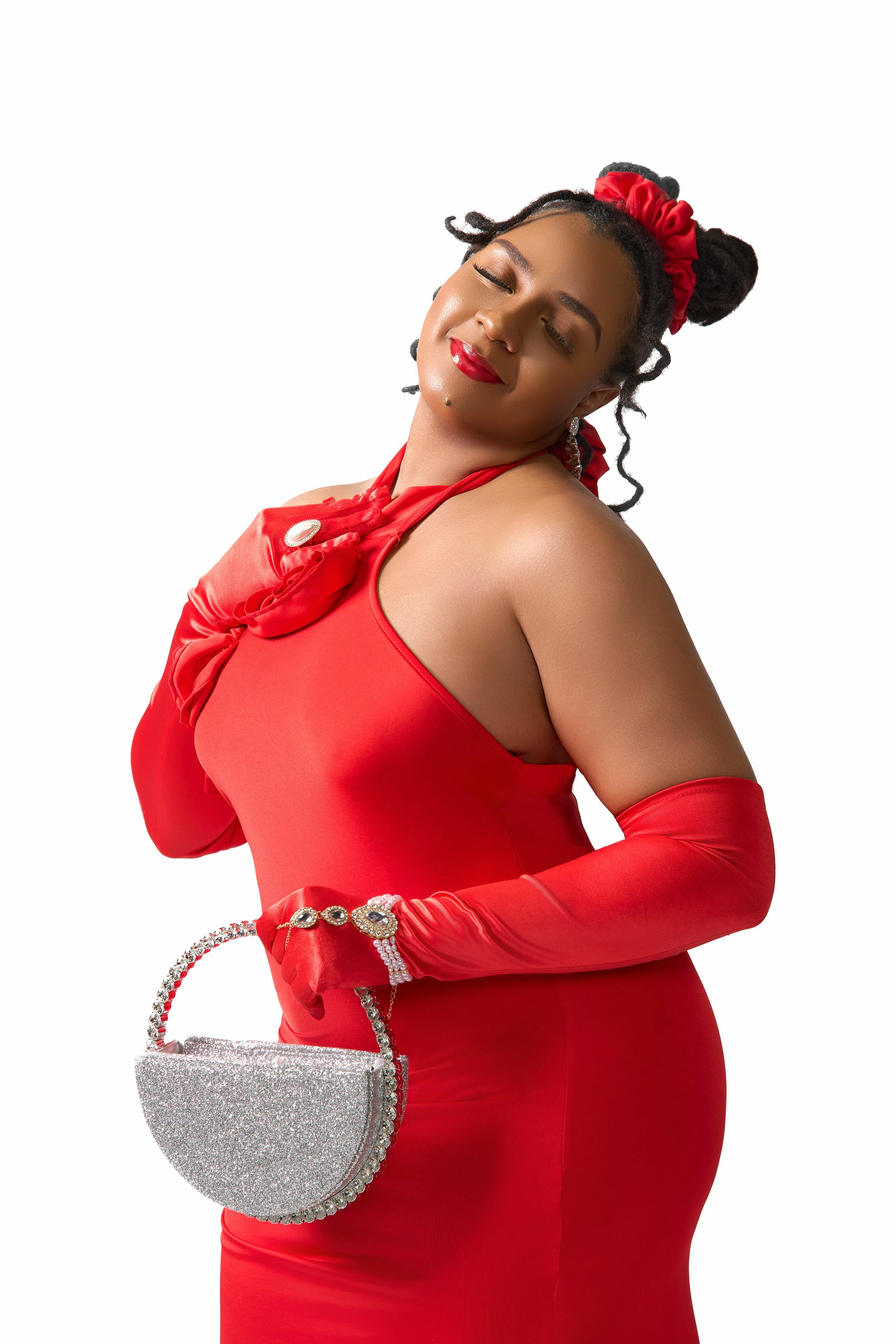 A woman dressed in a red gown with matching gloves, holding a glittery silver handbag, with her eyes closed and a content expression, against a plain white background.