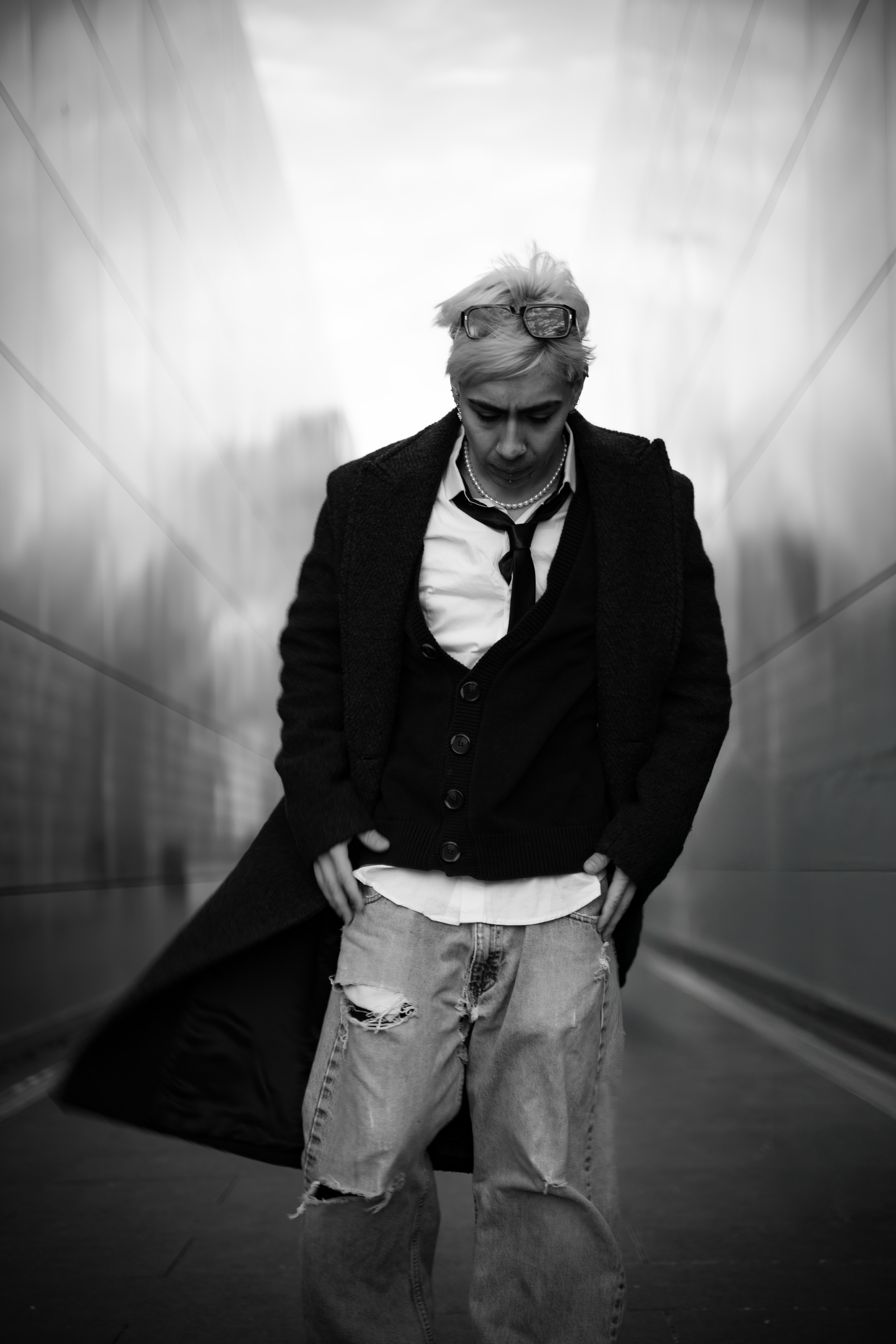 A person with short blonde hair, glasses on their head, wearing a white shirt, a black cardigan, and a dark coat, standing with their head bowed and hands in their pockets in an urban setting with metallic walls.