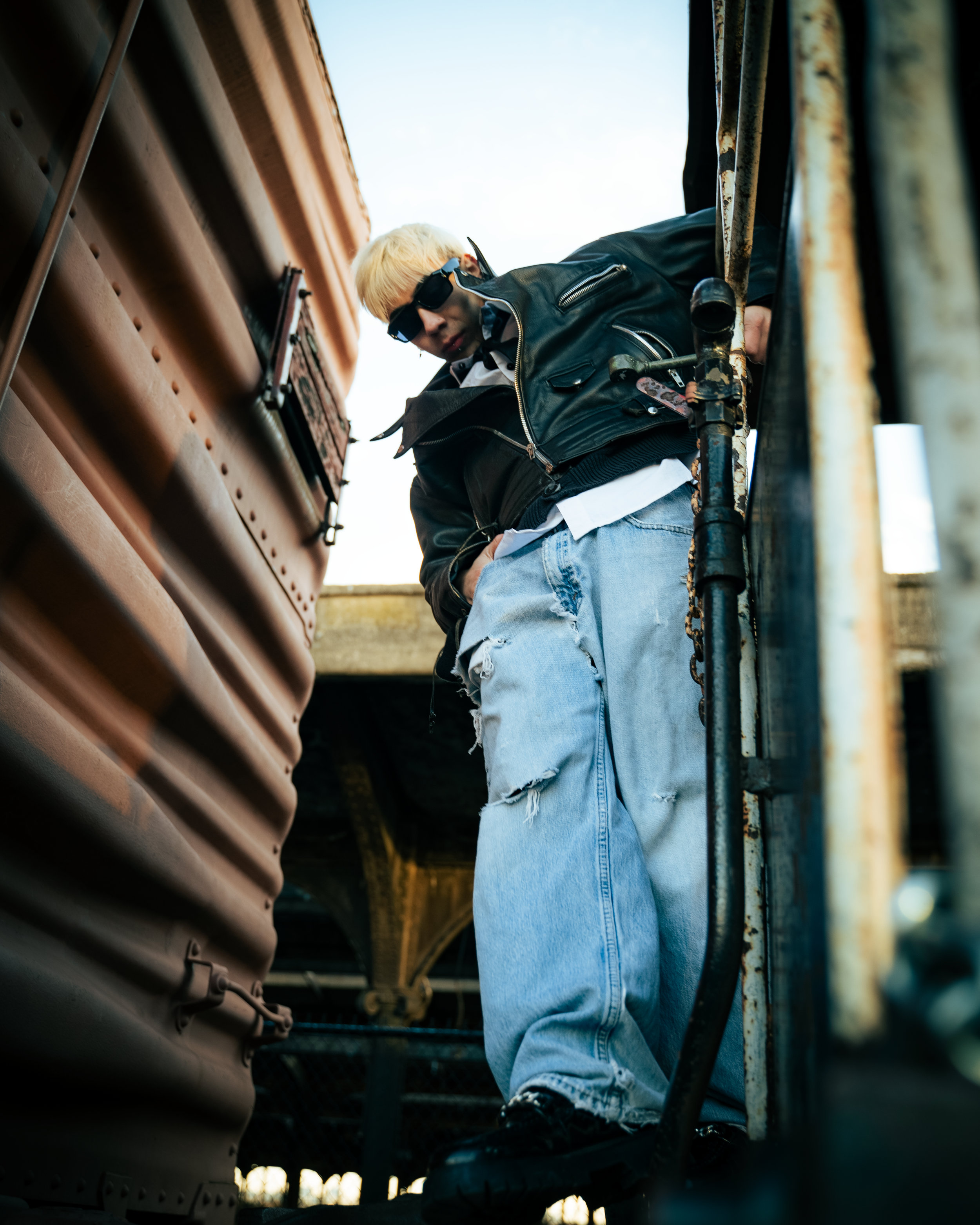 A young man with blonde hair, wearing sunglasses, a black leather jacket, and ripped jeans, standing on a rusty industrial staircase outdoors.