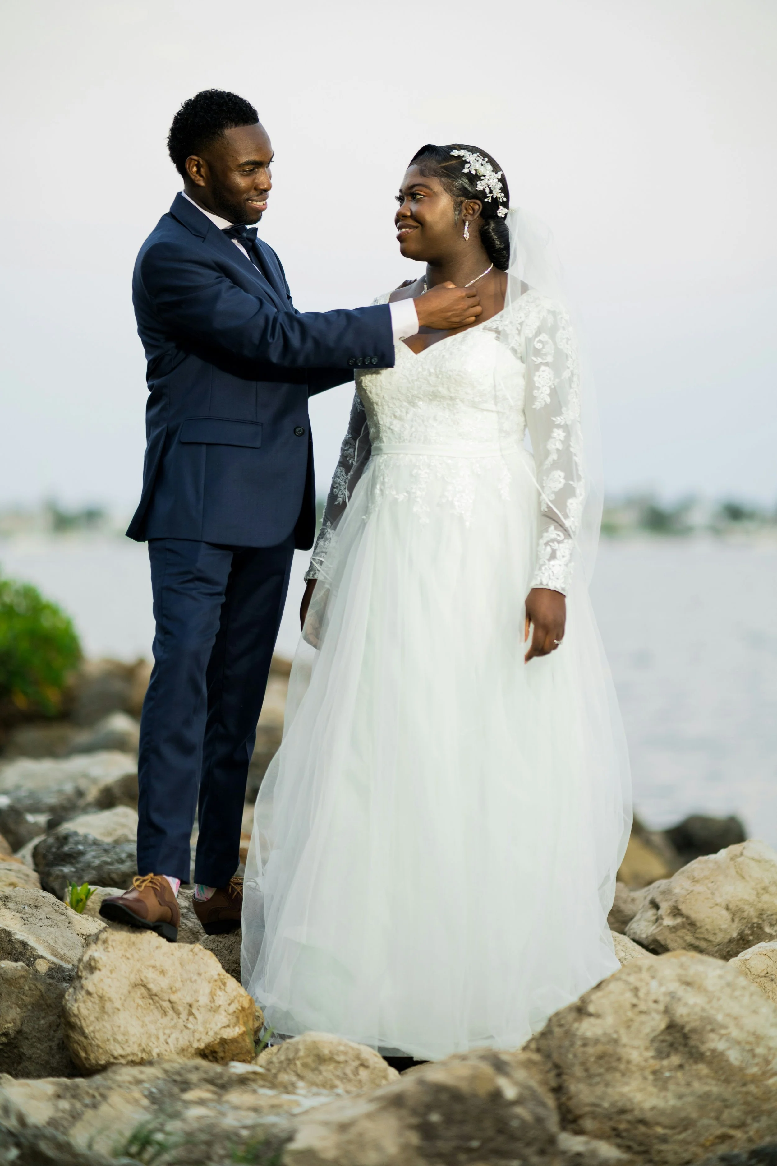 A couple on their wedding day standing on rocks by water, with the groom touching the bride's neck as they look at each other.