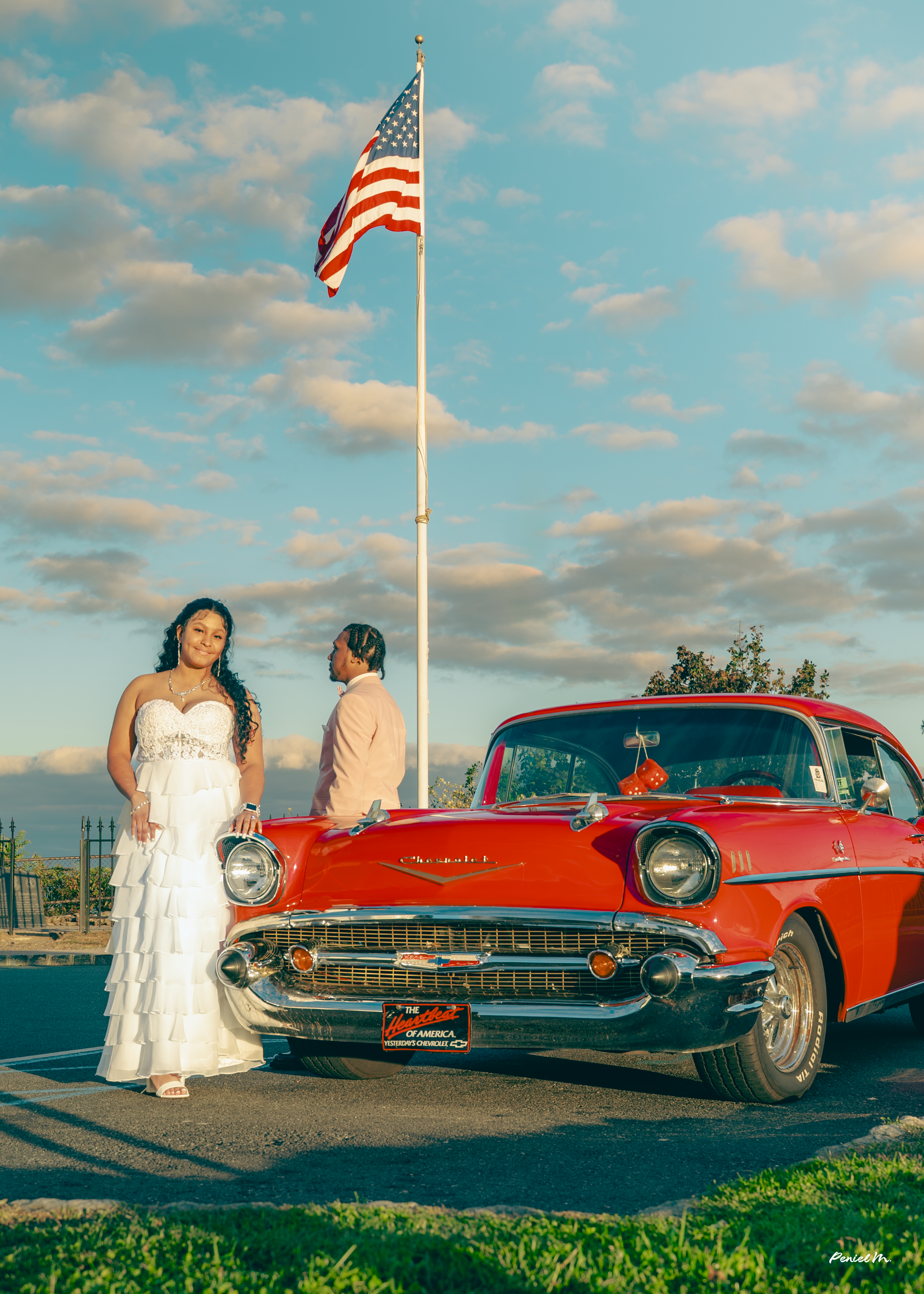 A woman in a white dress and a man in a light-colored suit stand next to a vintage red Chevrolet car with an American flag nearby, set against a partly cloudy sky during the daytime.
