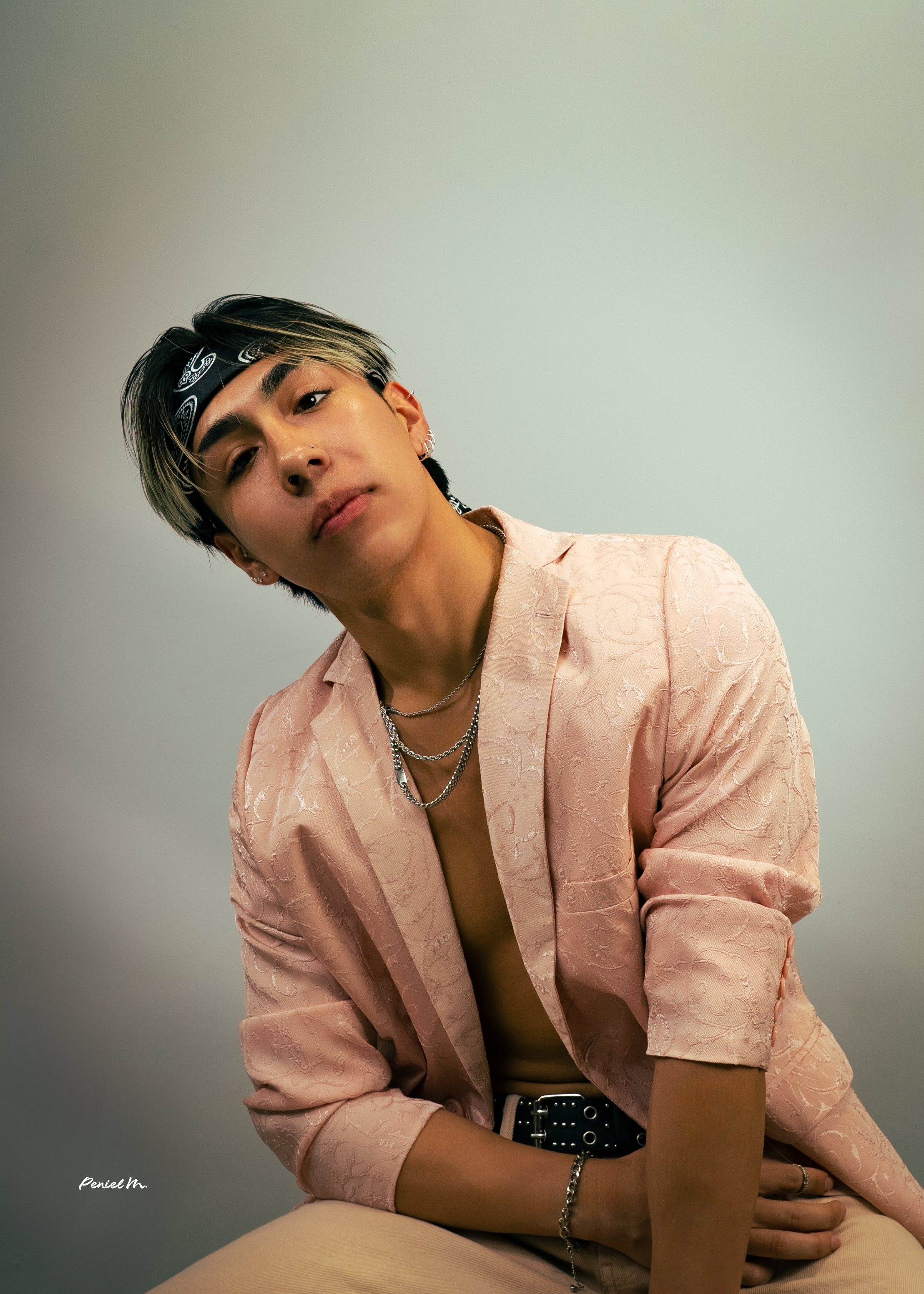 A young man with black and blond hair, wearing a black bandana, layered necklaces, and a pink patterned blazer, poses against a plain background.