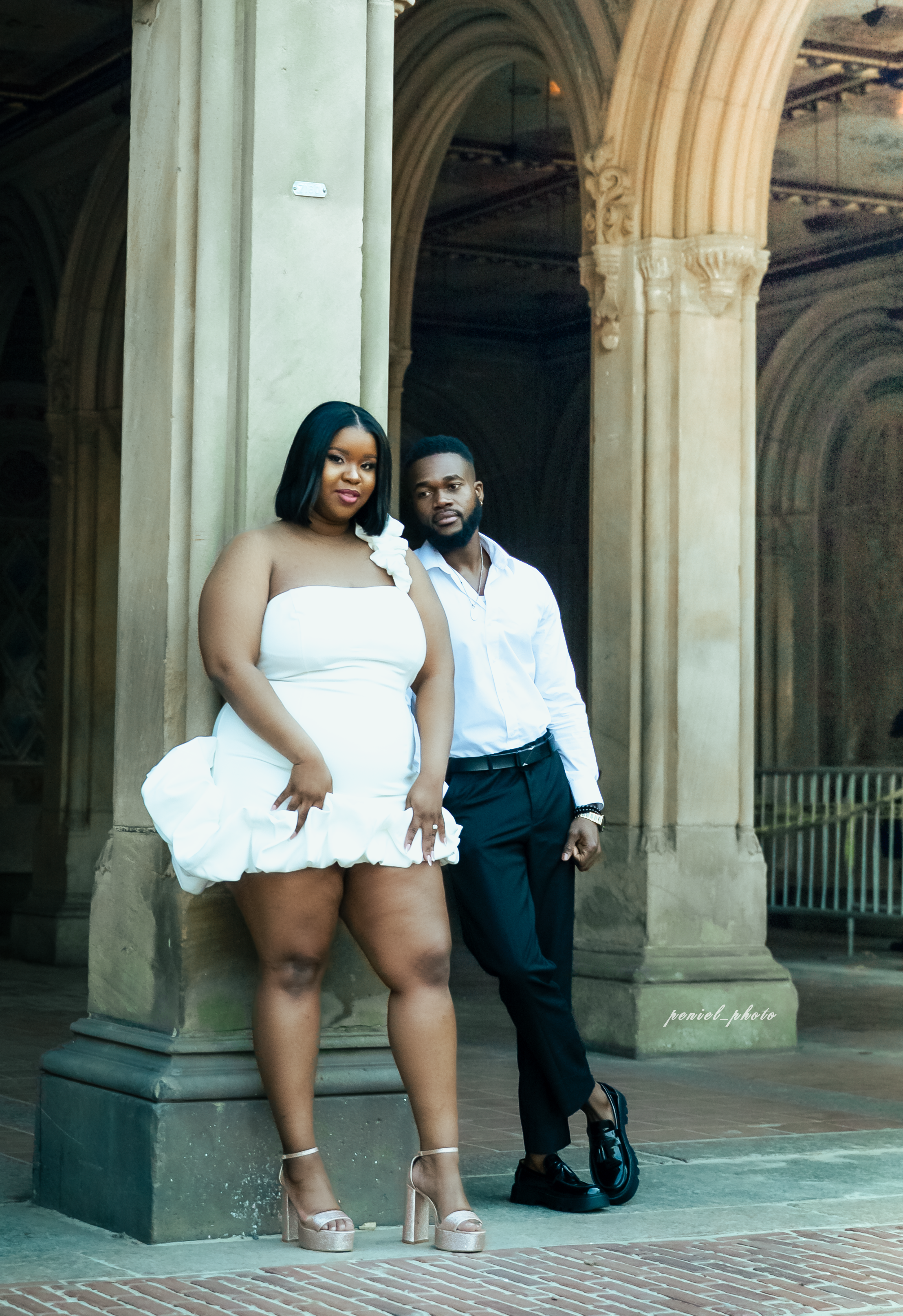 A woman and man standing inside a historic arched building, with the woman in a white strapless dress and high heels, and the man in a white shirt, black pants, and dress shoes, posing together.