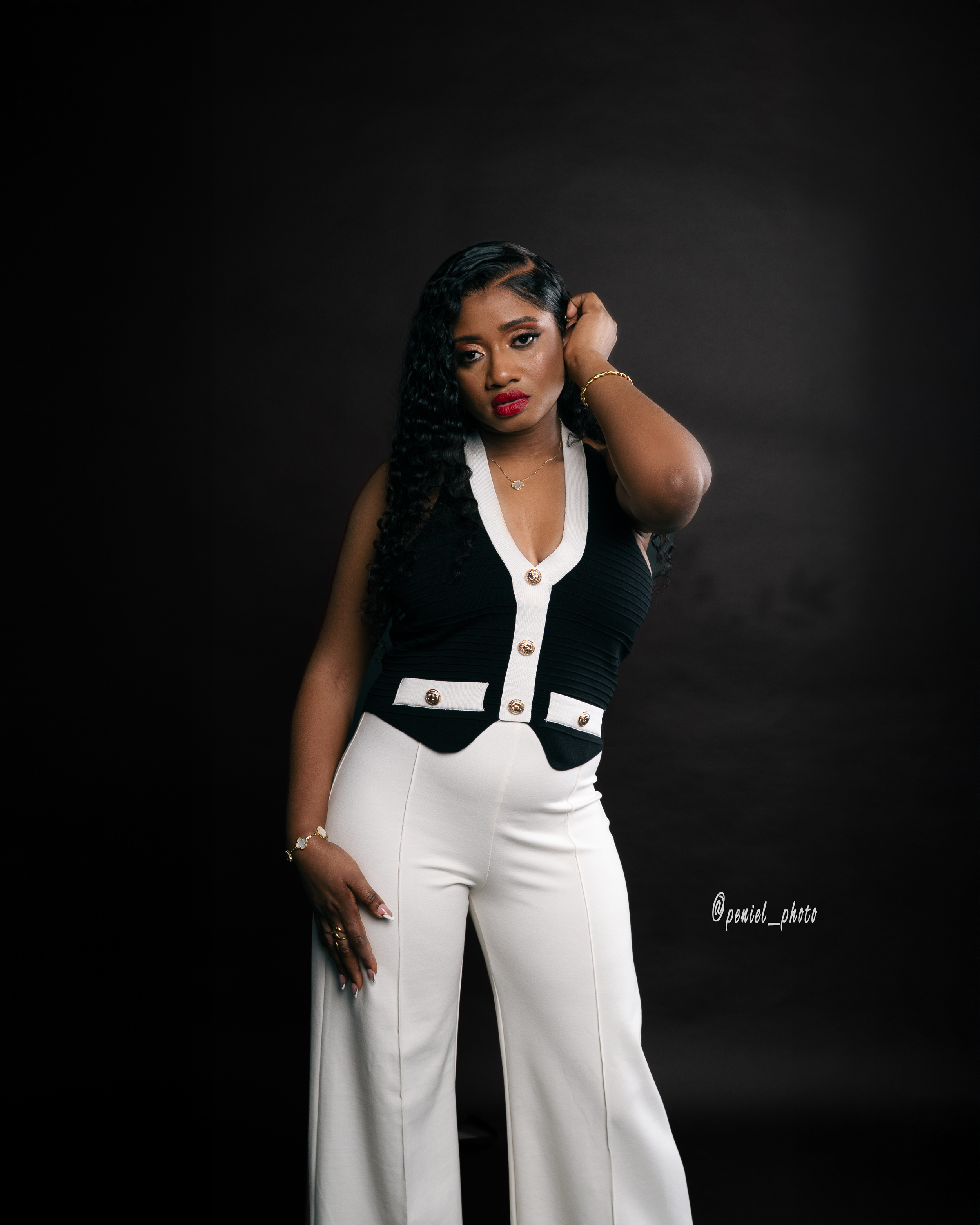 A woman with long, curly black hair and red lipstick poses against a dark background, wearing a black and white sleeveless top with gold buttons and white wide-leg pants.