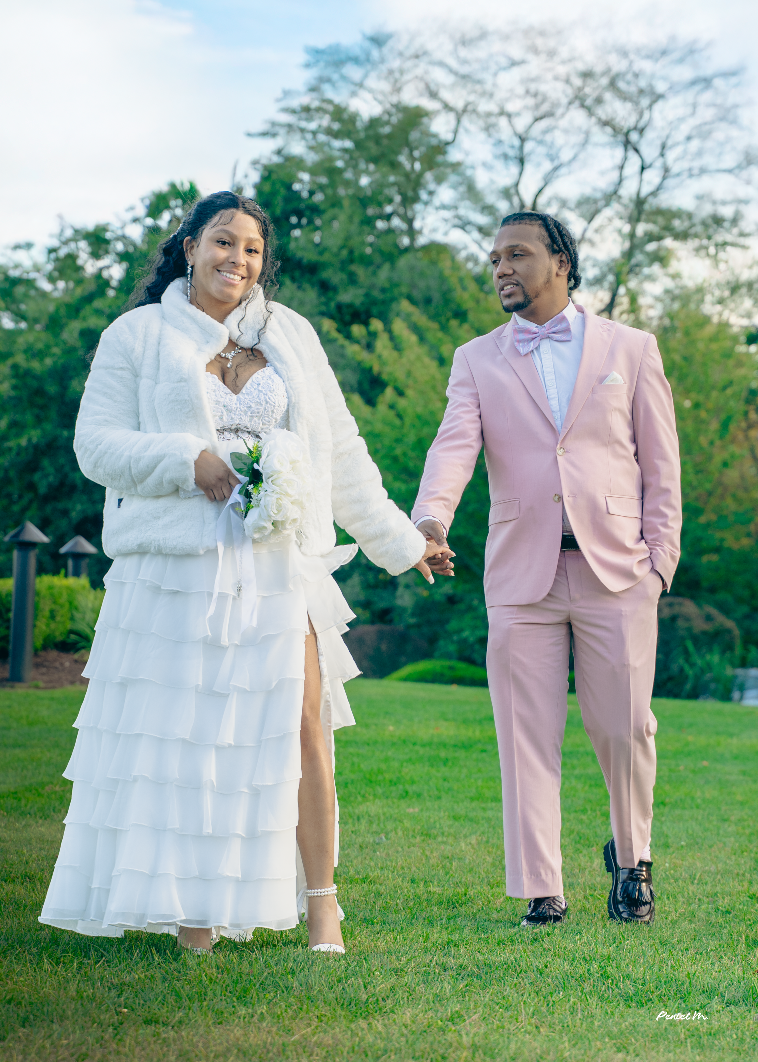 A bride and groom holding hands outdoors on a grassy area with trees in the background, dressed in wedding attire, smiling and looking happy.