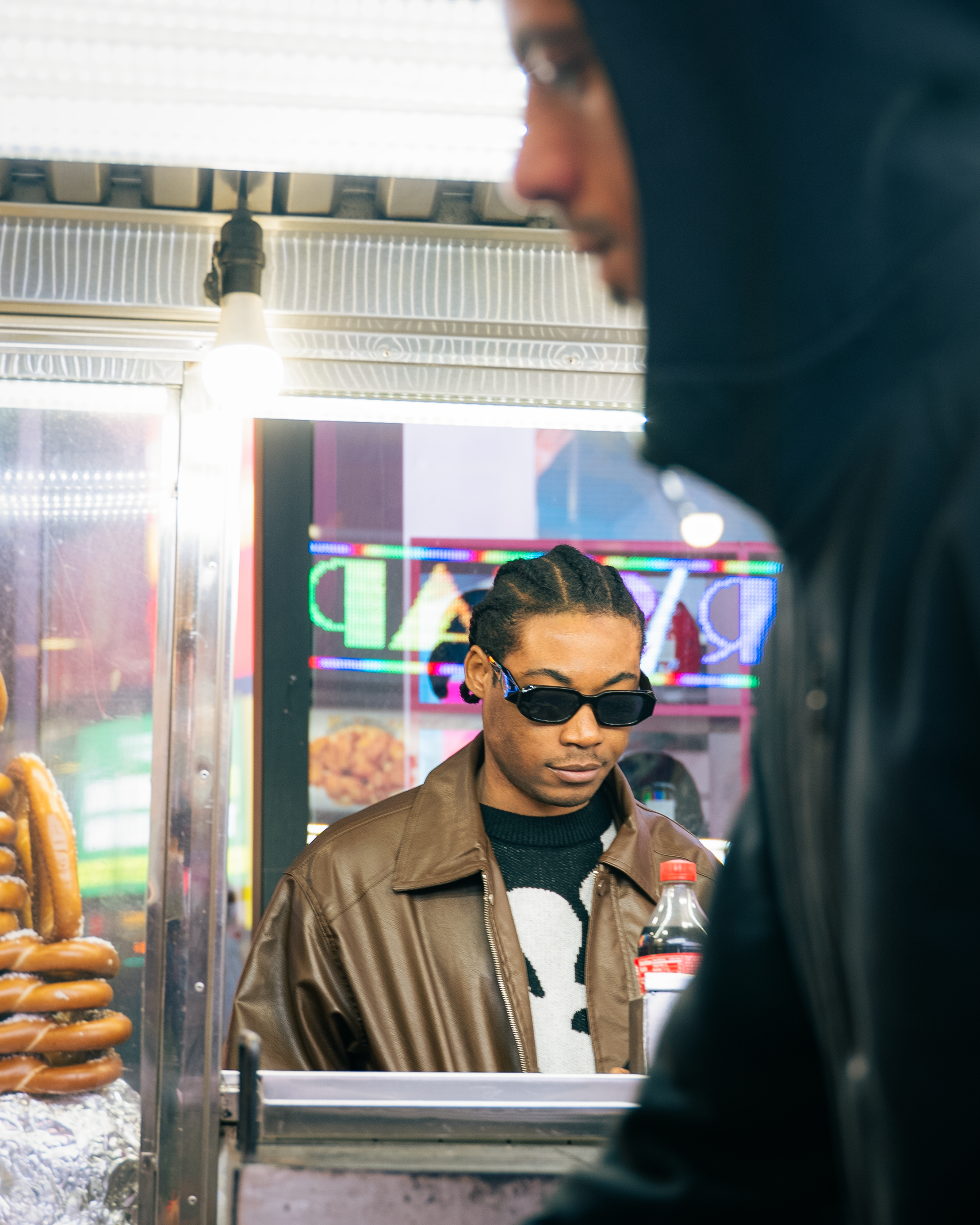 A person with dreadlocks and sunglasses working at a food stand with hot dogs on display. Another person in a black jacket is in the foreground.