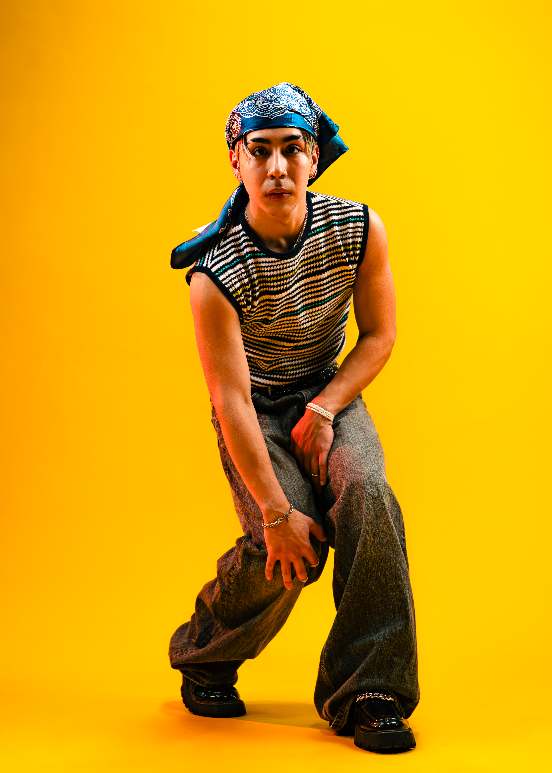 Person crouching in front of a yellow background, wearing a sleeveless striped shirt, baggy pants, a blue bandana, and chunky black shoes.