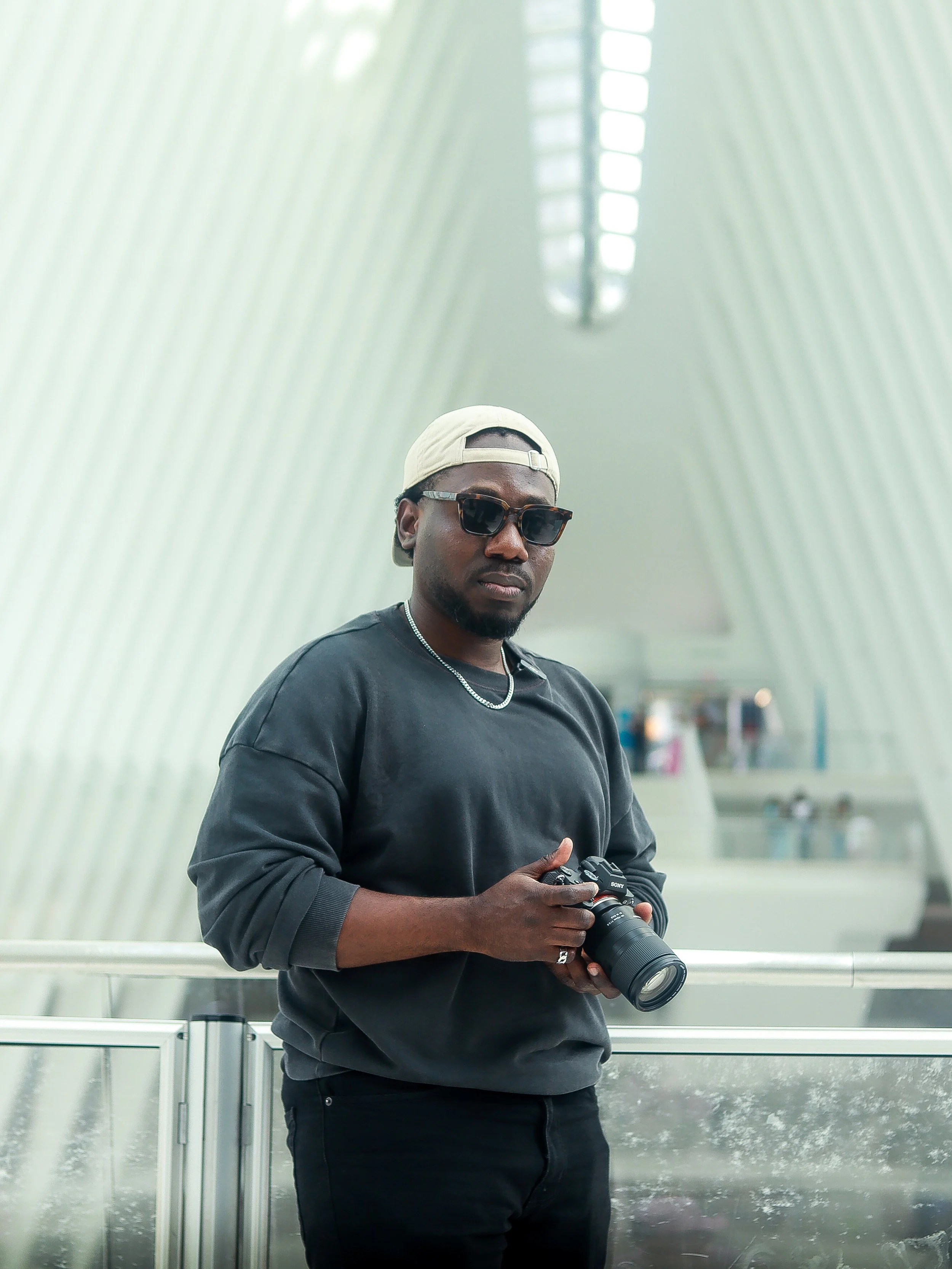 A young man wearing sunglasses, a beige baseball cap backwards, a black sweatshirt, and a silver chain holds a DSLR camera. He stands in an indoor space with a modern, curved, light-colored architectural background.