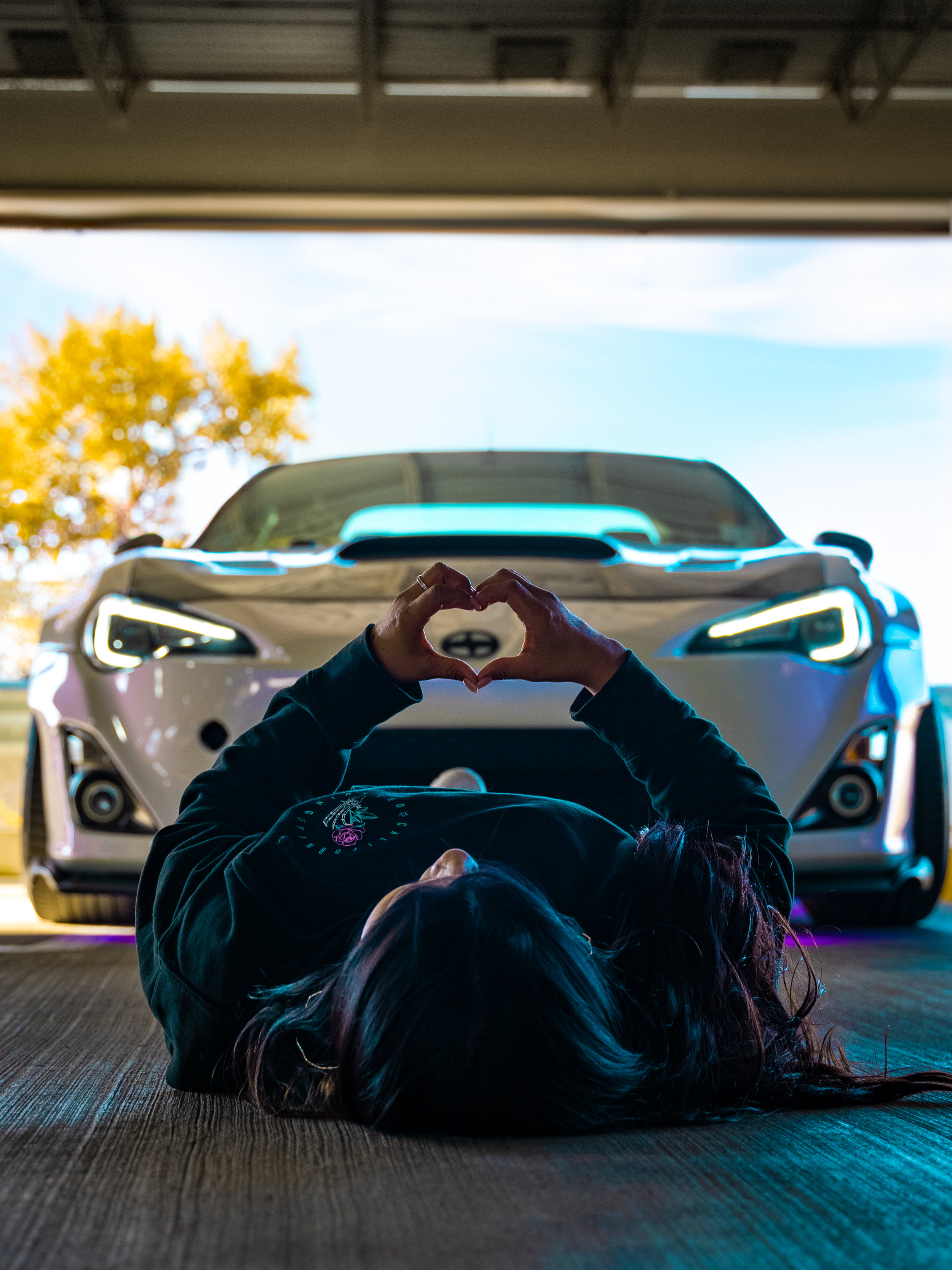 Person lying on the ground in front of a white car making a heart shape with their hands.