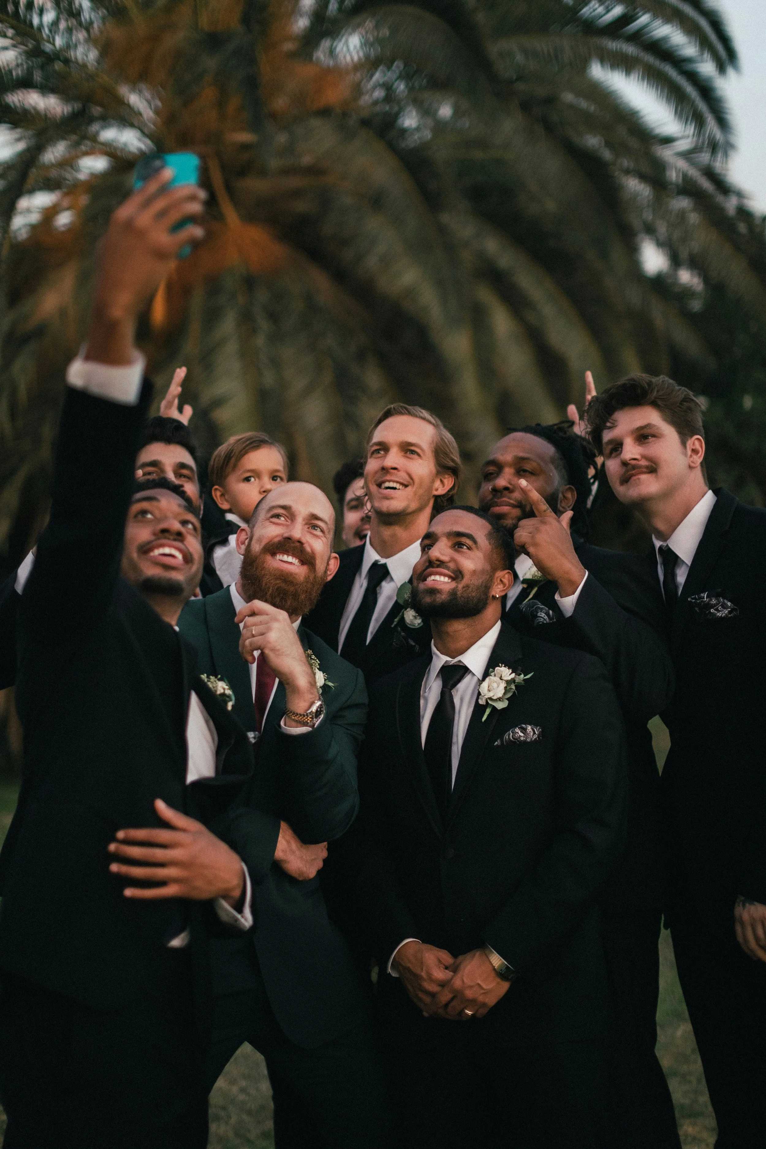 Group of men dressed in formal suits taking a selfie outdoors with palm trees in the background.