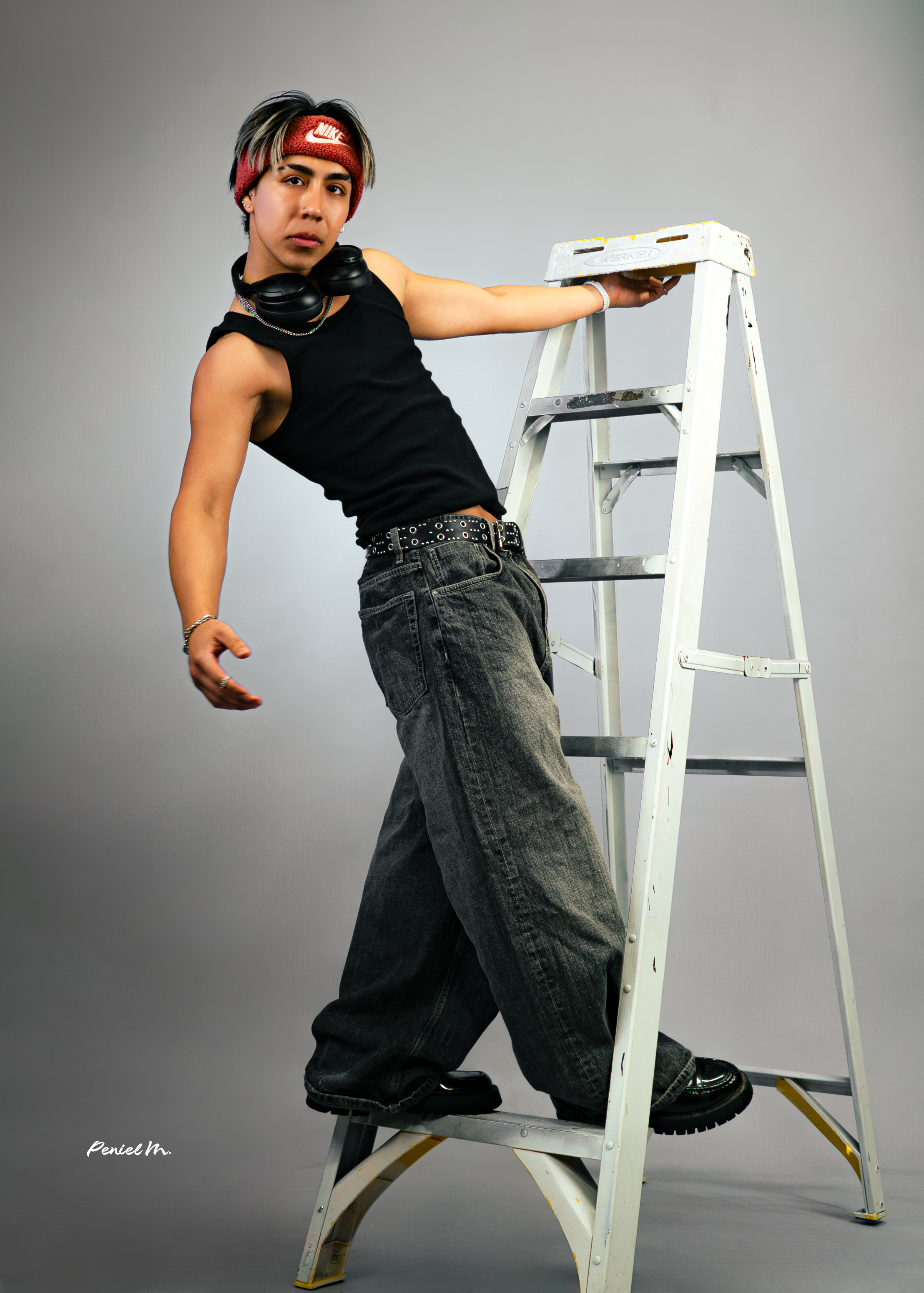 Person standing on a ladder with one hand resting on the top step, wearing a red headband, big headphones around neck, black sleeveless shirt, black baggy jeans, and black shoes, against a plain grey background.