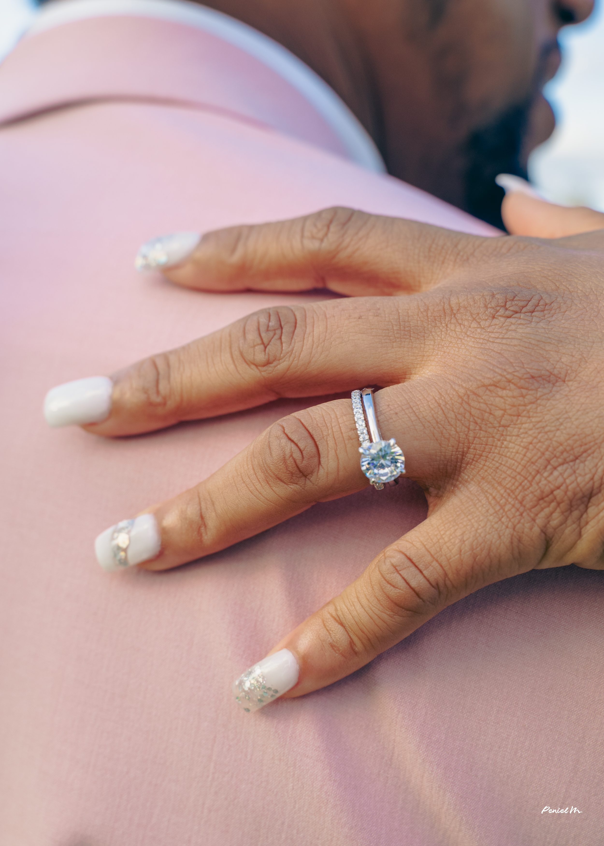 Close-up of a person displaying a diamond engagement ring on their ring finger, with well-maintained nails and a pink garment in the background.