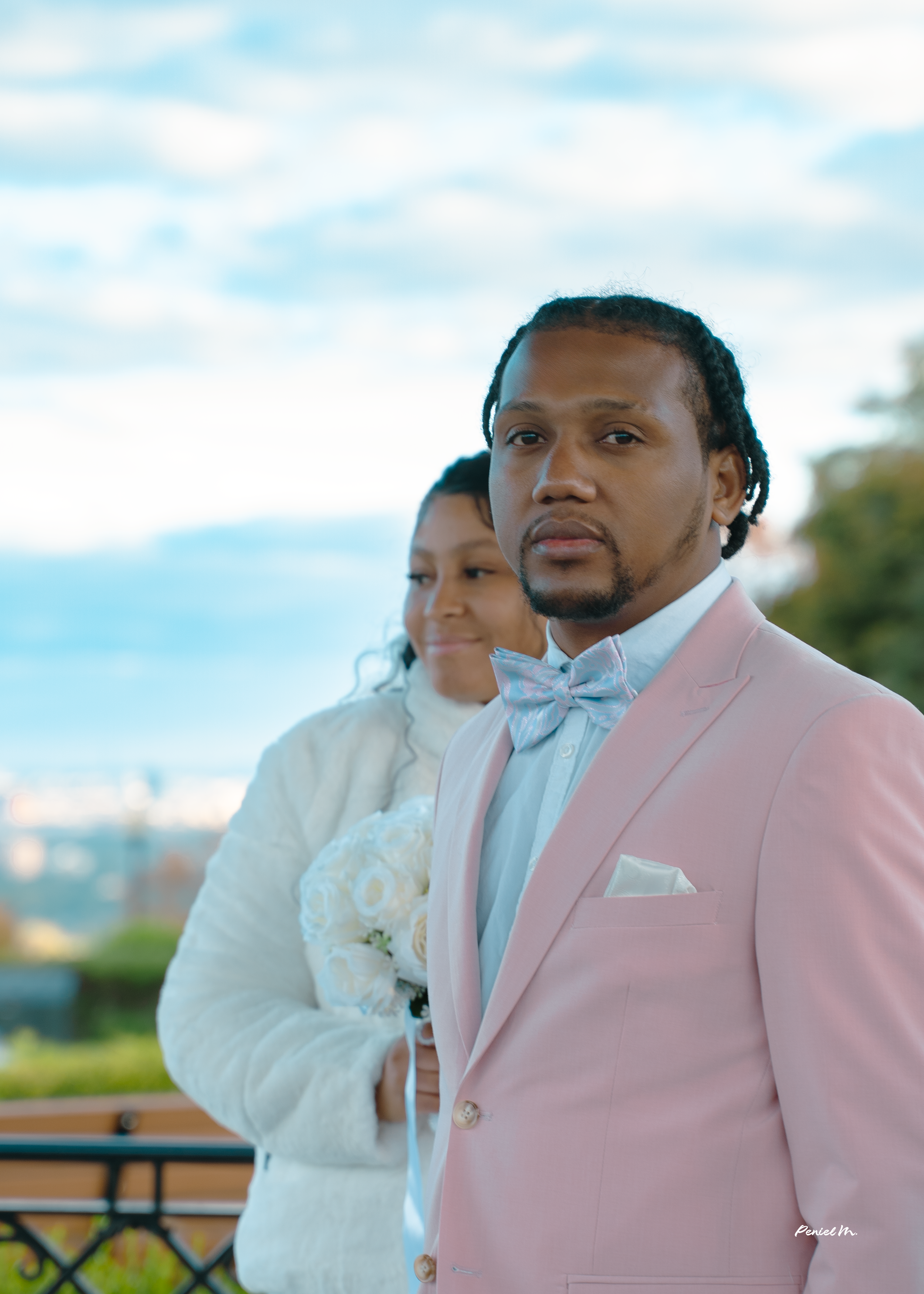 A man in a pink suit with a lavender bow tie and a woman in a white dress holding a bouquet of white roses, standing outdoors with a cityscape and sky in the background.